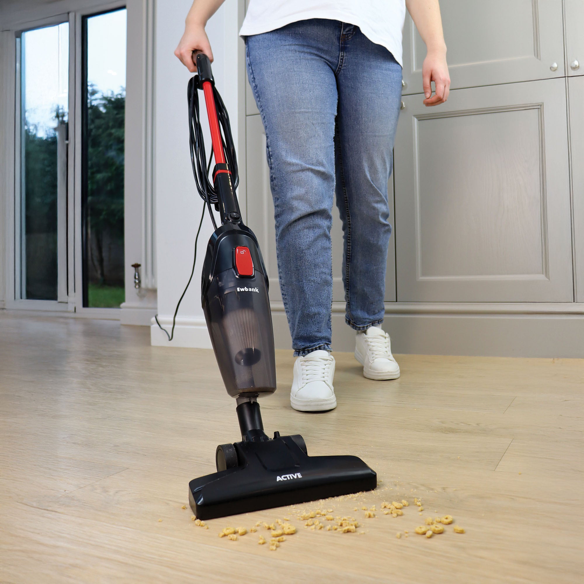 Wearing jeans and white shoes, a person uses the ACTIVE 2-in-1 Corded Stick Vacuum Cleaner in red/black, featuring a washable filter, to clean crumbs from a light wood floor in a modern kitchen.