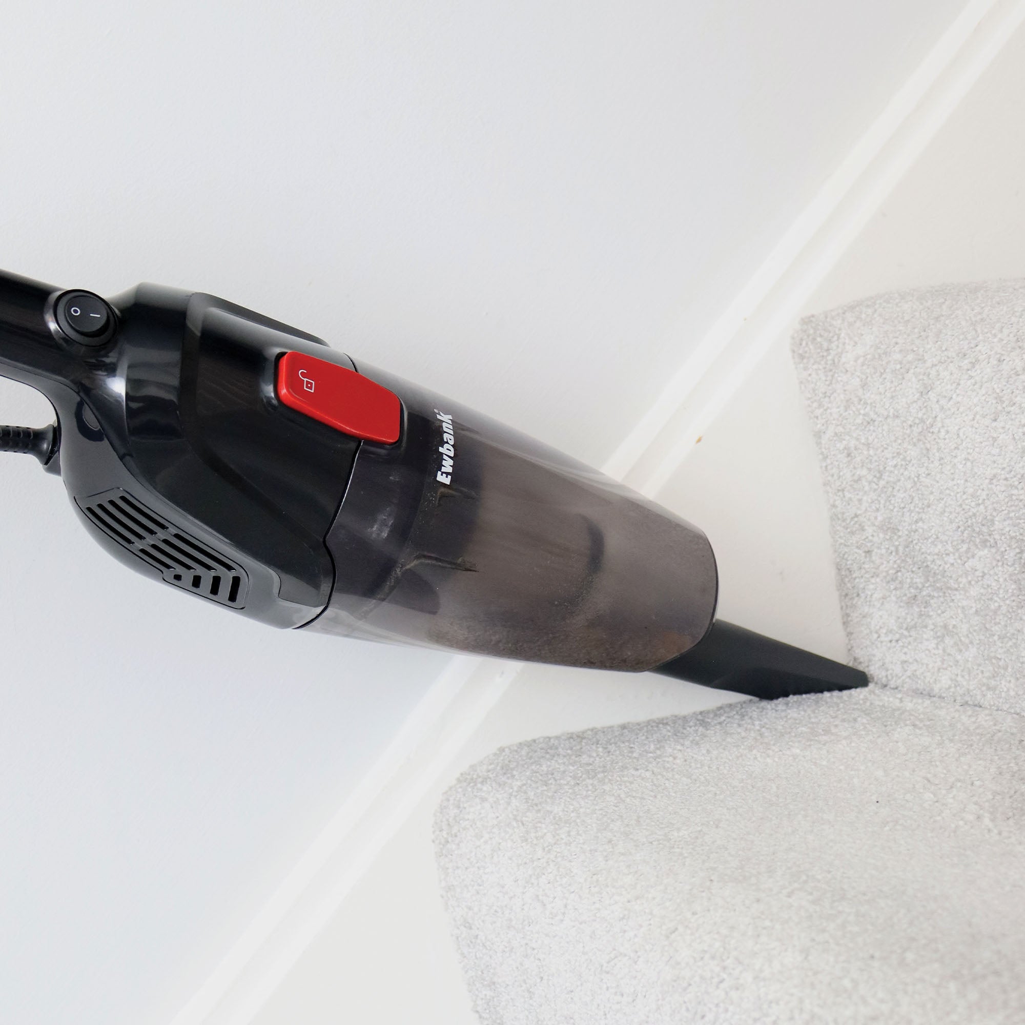 The ACTIVE 2-in-1 Corded Stick Vacuum Cleaner in red/black, featuring a washable filter, is being used to clean the corner of a room with light-colored carpet and white walls.