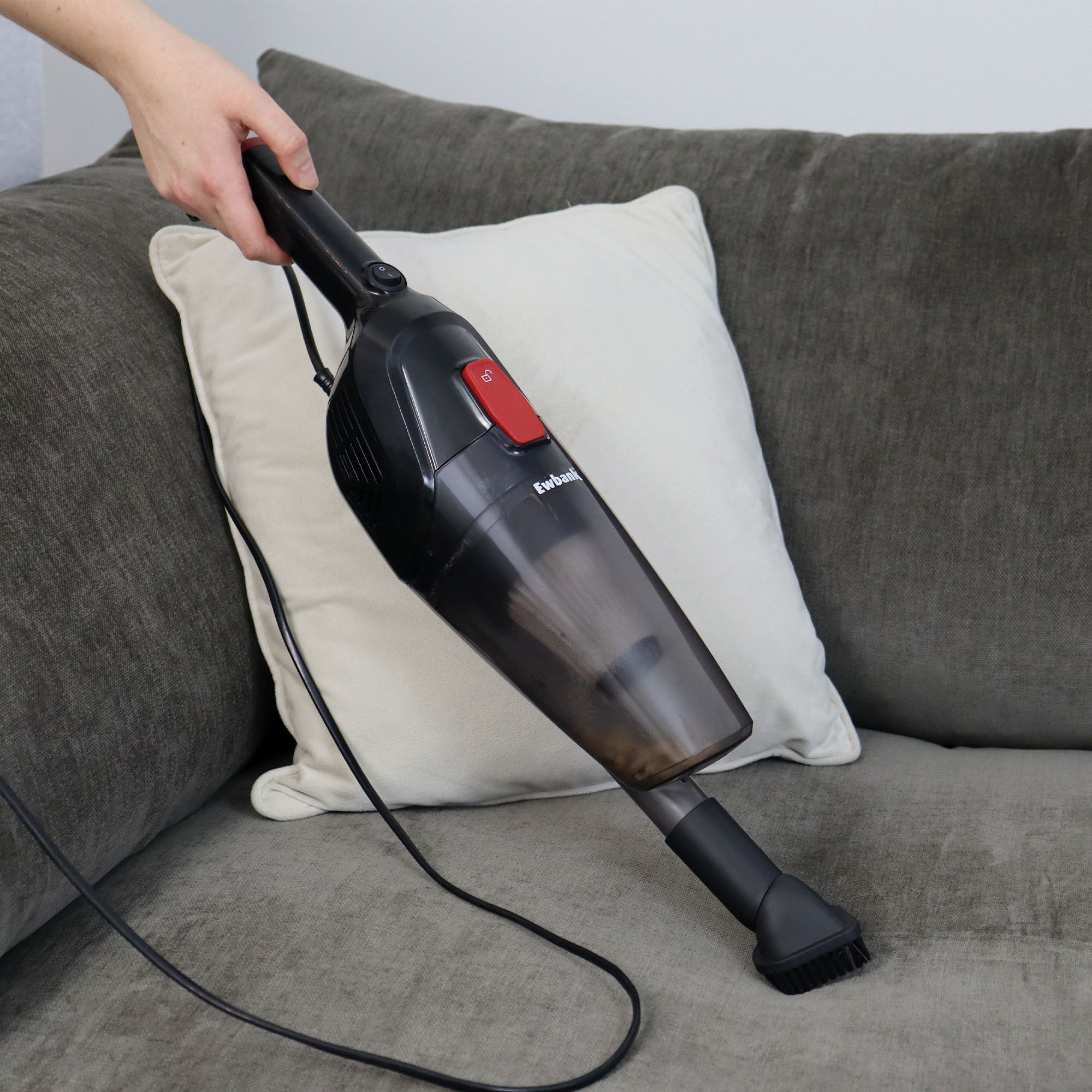 A person cleans a dark gray couch with a light-colored pillow using the ACTIVE 2-in-1 Corded Stick Vacuum Cleaner, Red/Black, which is lightweight and includes a brush attachment and washable filter.