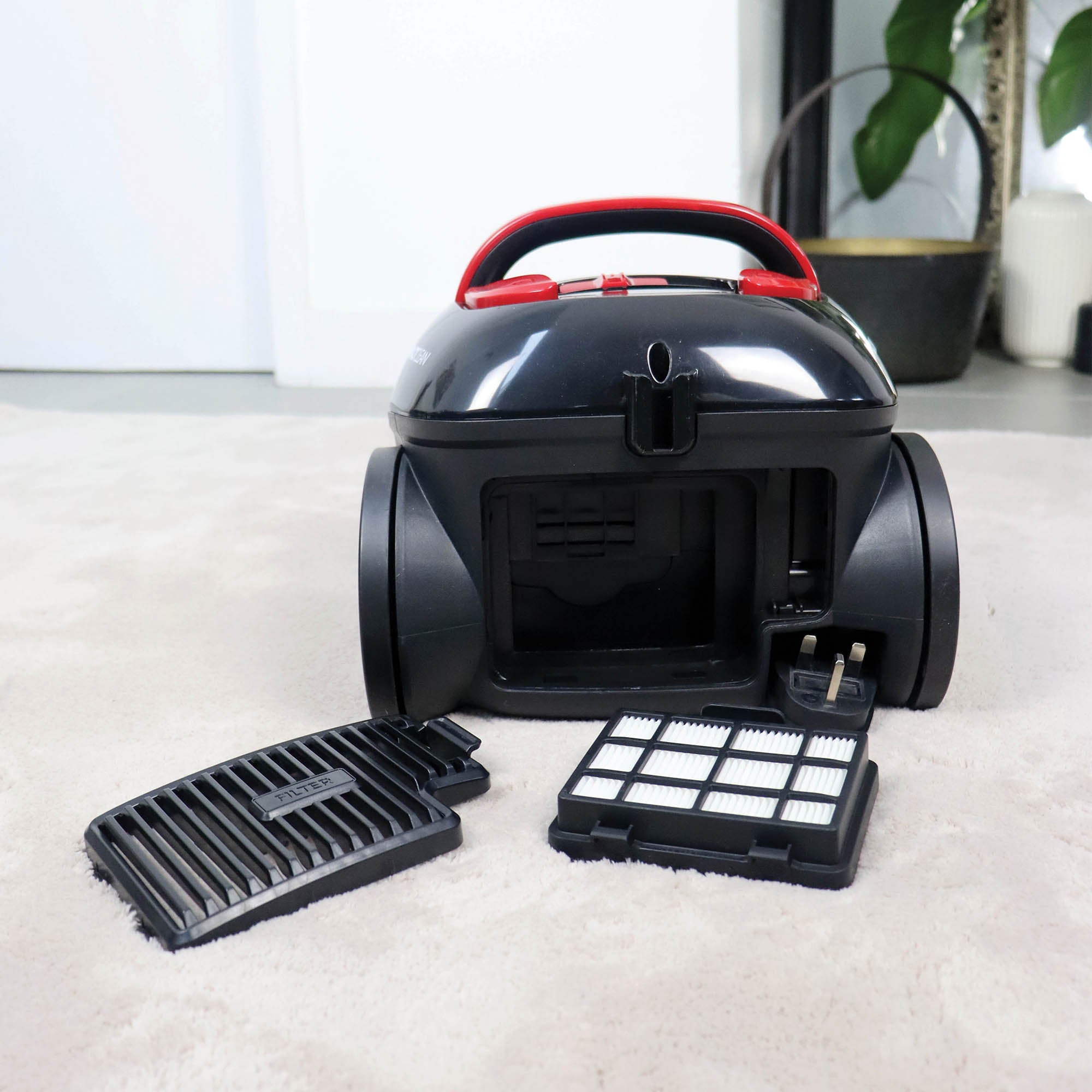 A Ewbank COMPACTCLEAN Bagged Vacuum Cleaner in black and red sits on a beige carpet with its back panel open, displaying the removed HEPA filter and grille. A green potted plant appears in the background.