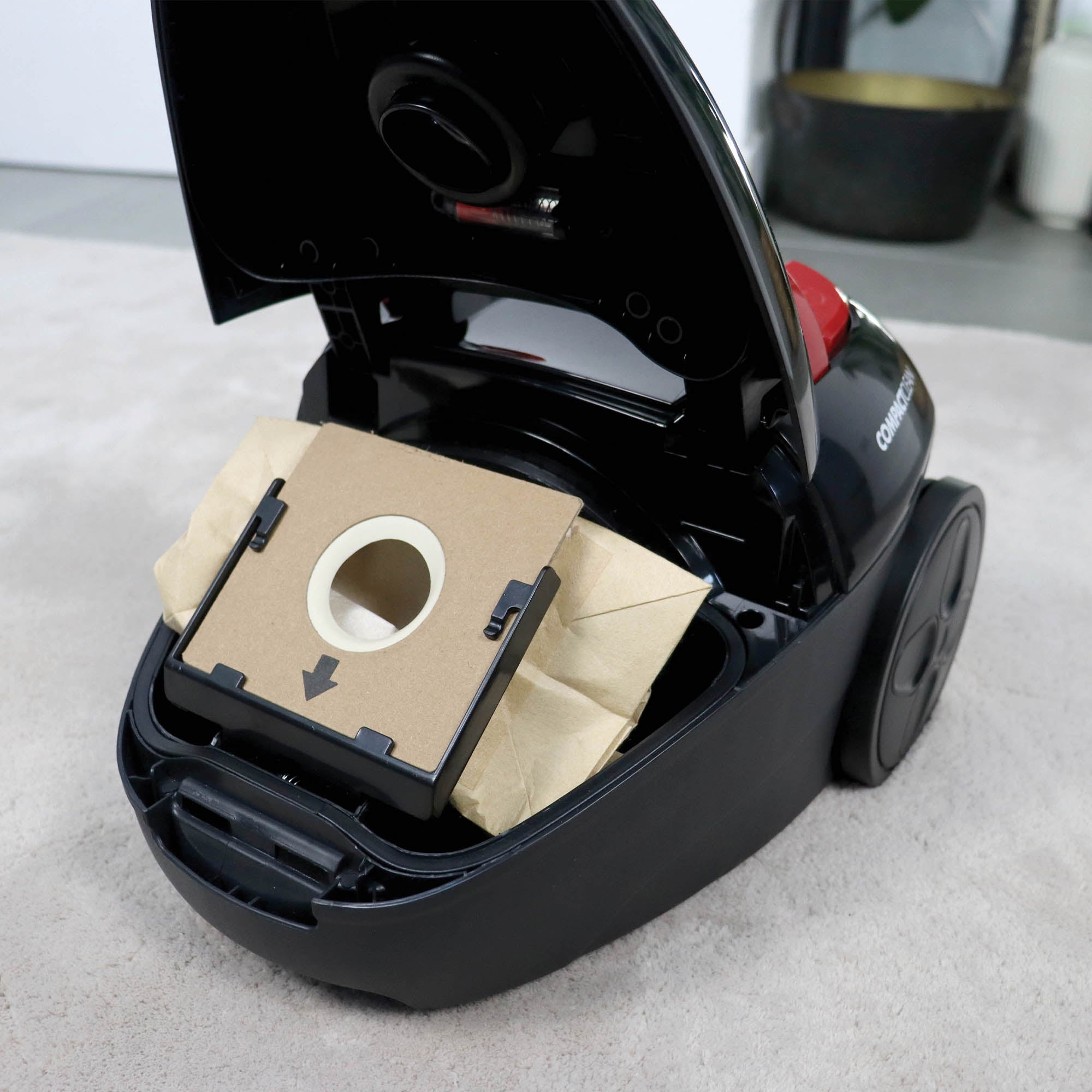 A close-up shows the Ewbank COMPACTCLEAN Bagged Vacuum Cleaner open on a light-colored carpet, revealing its replaceable dust bag inside the compartment.