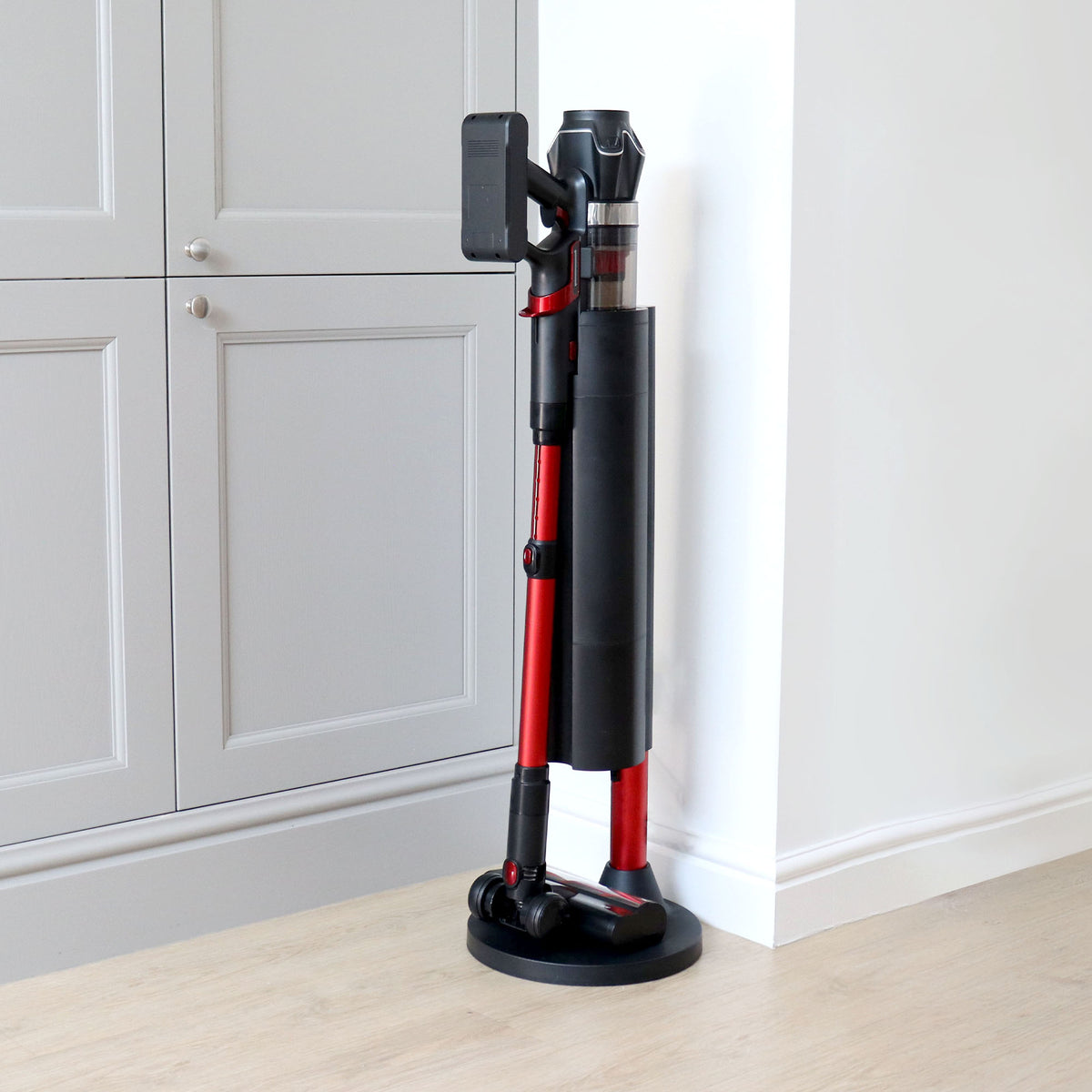 The AIRSTORM AUTO Empty Cordless Stick Vacuum Cleaner, in red and black, stands upright on its charging base with an auto-emptying dock in the corner of a room featuring light wood flooring and gray cabinets.