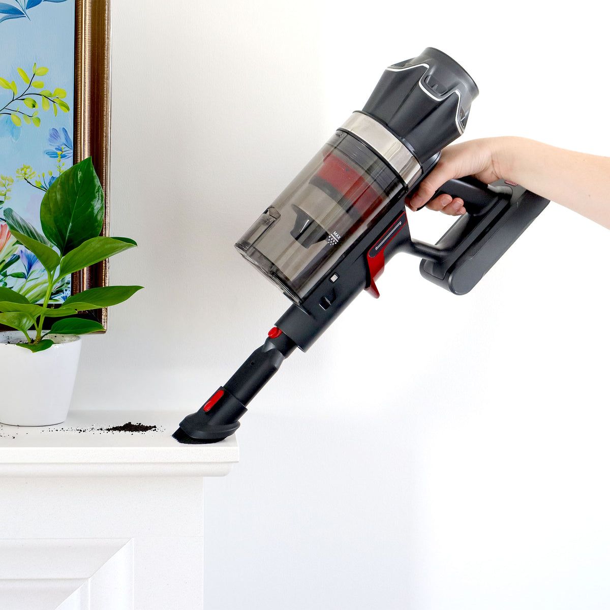 A hand holds the AIRSTORM AUTO Empty Cordless Stick Vacuum Cleaner, using its handheld mode to clean spilled soil from a white shelf beside a plant and a colorful framed picture.