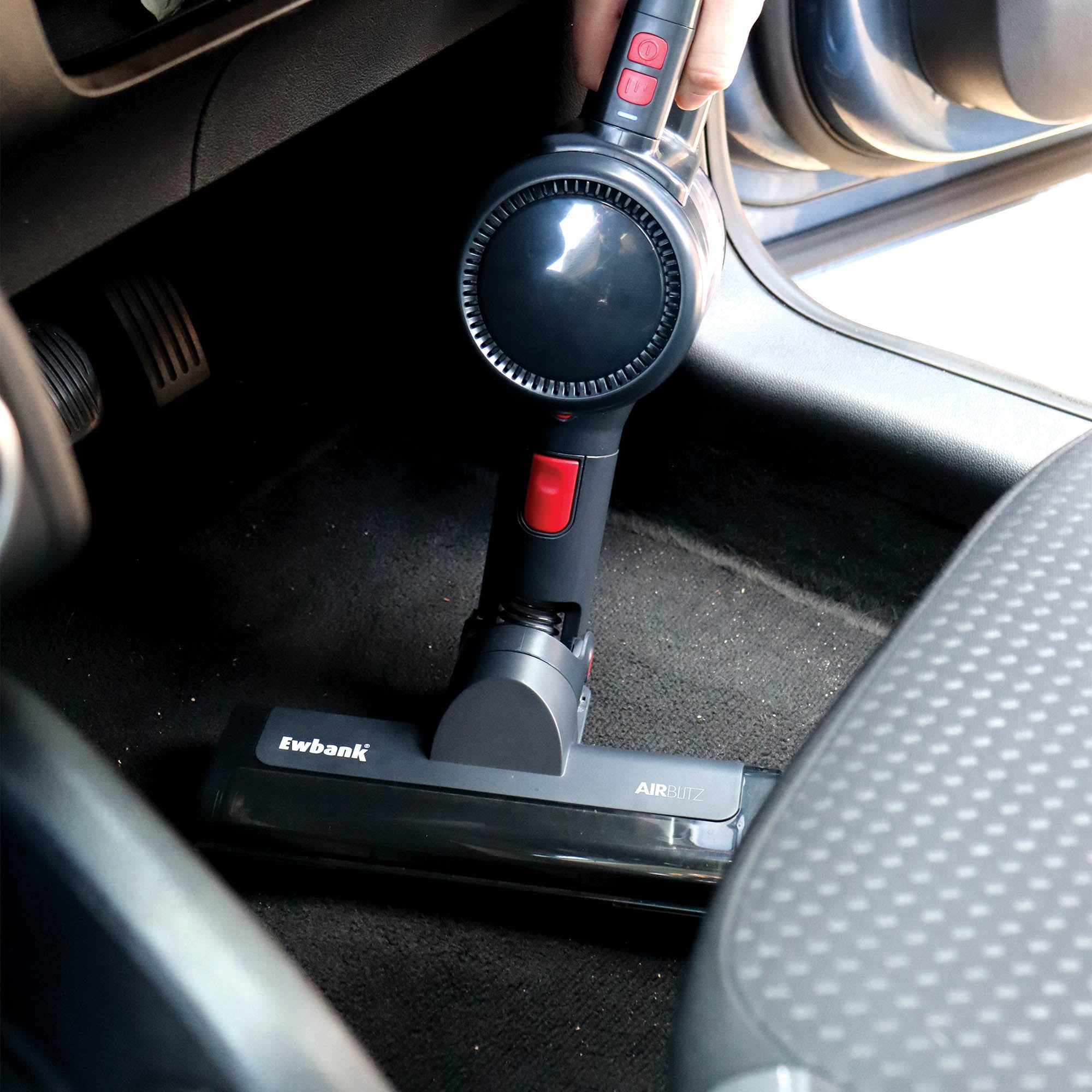 A person uses the AIRBLITZ 2-in-1 Cordless Stick Vacuum Cleaner with a washable HEPA filter to clean the black carpet floor near the driver’s seat inside a car.