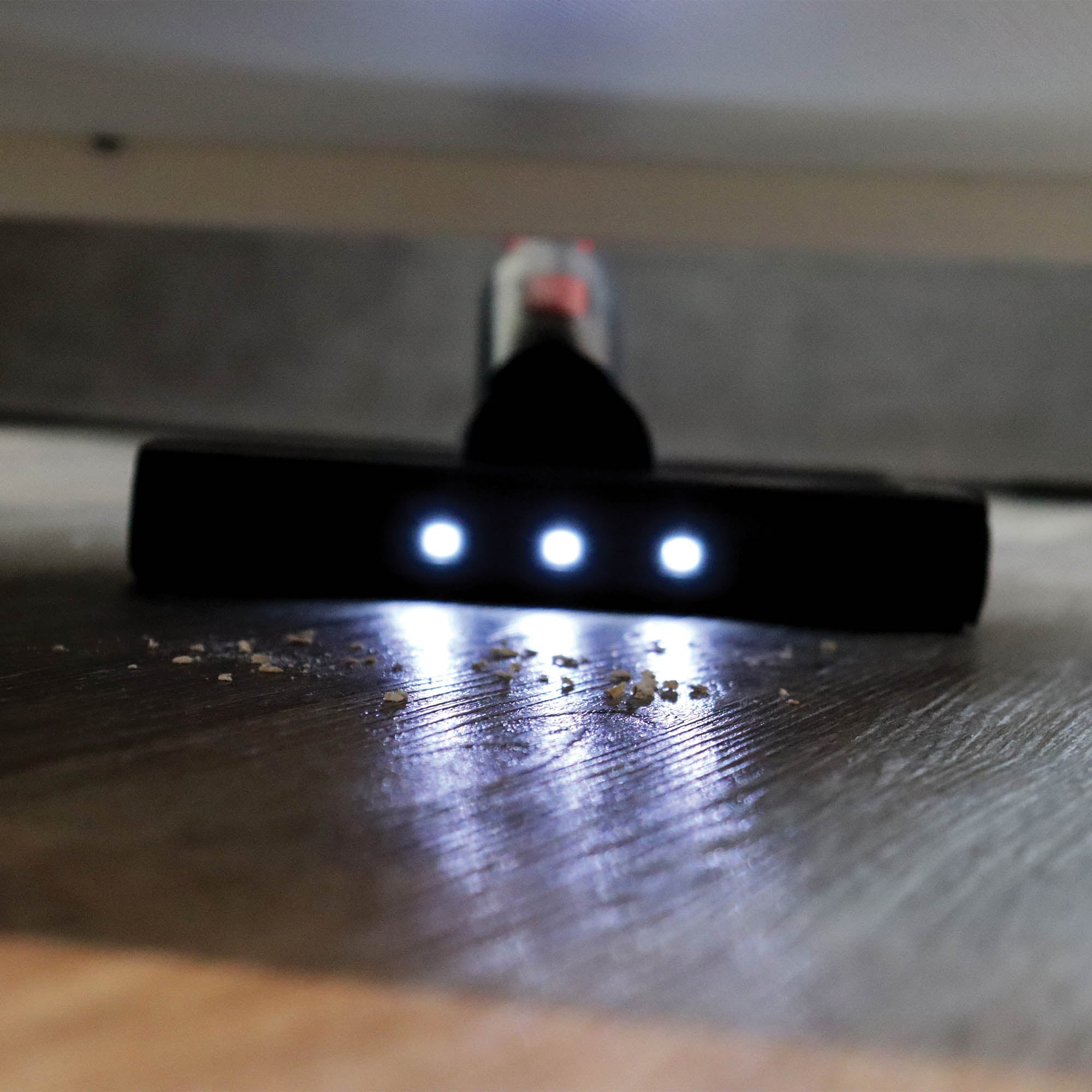 Close-up of the AIRBLITZ 2-in-1 Cordless Stick Vacuum Cleaner head with three white LED lights, cleaning crumbs on a hardwood floor beneath furniture.