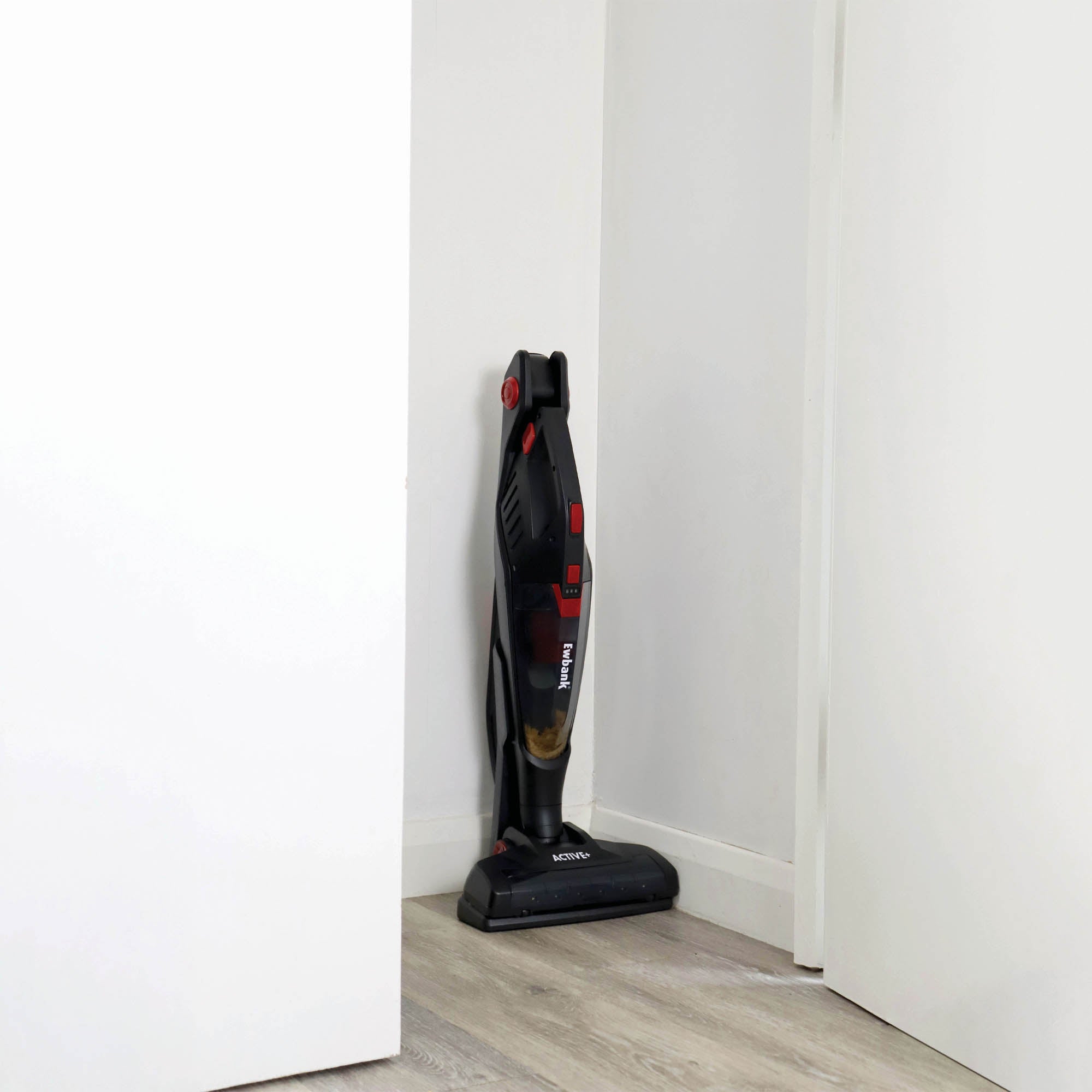The ACTIVE+ 2-in-1 Cordless Stick Vacuum Cleaner, in black and red with a HEPA filter, stands upright in the corner of a room with white walls and light wooden floors, positioned between two open doors.