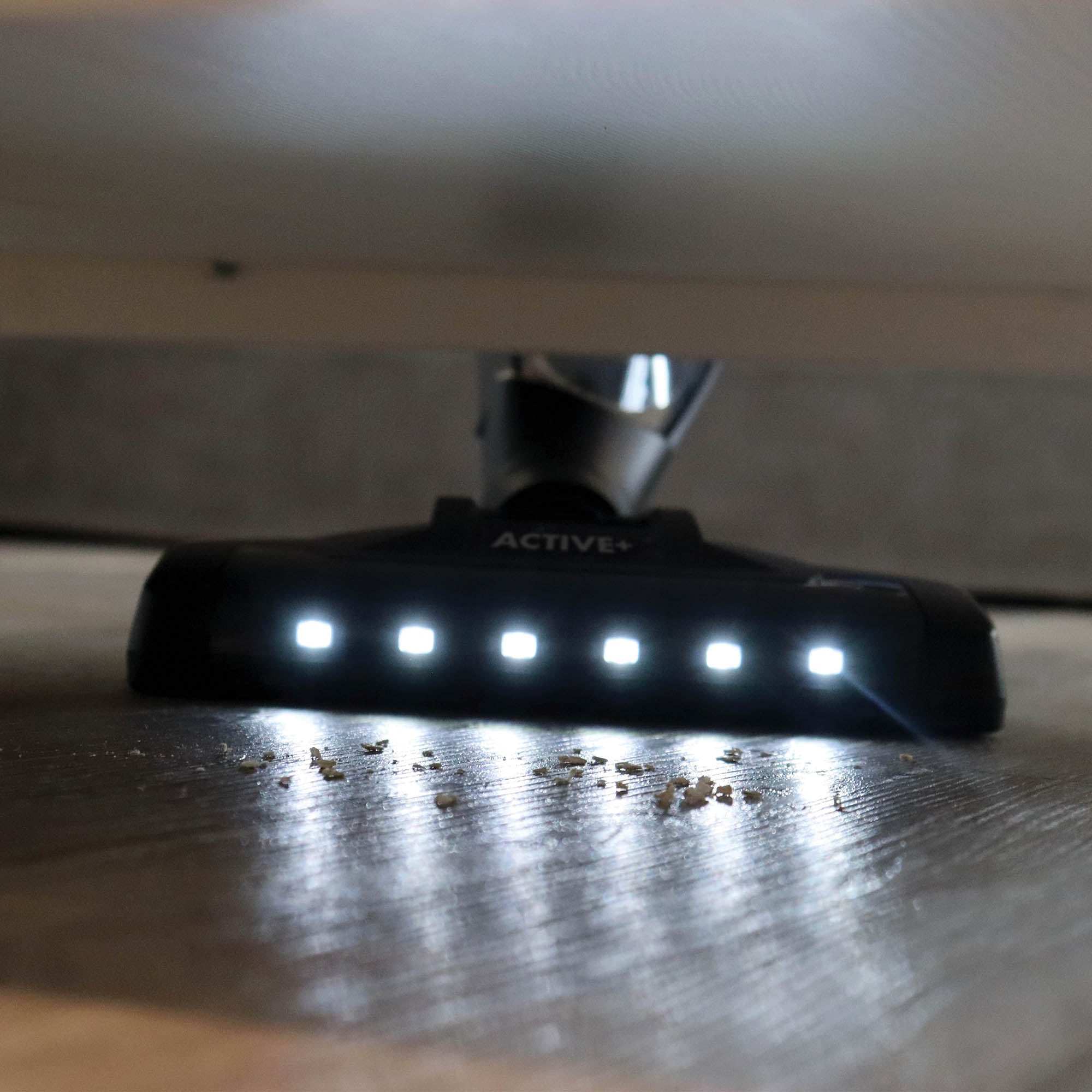 The ACTIVE+ 2-in-1 Cordless Stick Vacuum Cleaner, equipped with LED lights, cleans small debris from a hardwood floor beneath furniture.