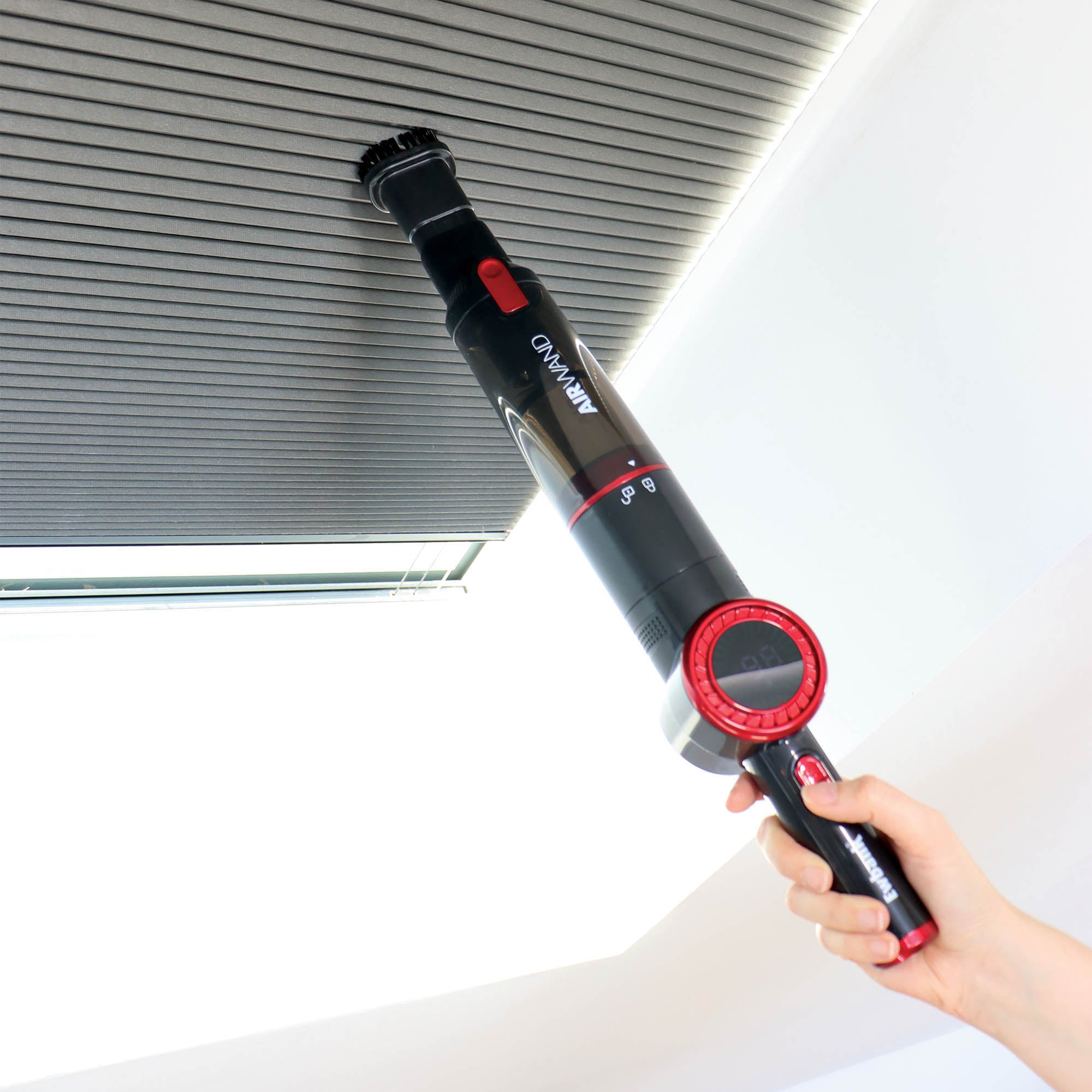 A hand uses the AIRWAND Cordless Handheld Vacuum Cleaner with a brush attachment to remove dust from a ceiling vent's grooves. The lightweight, black and red device is cordless and perfect for above-the-floor cleaning tasks.