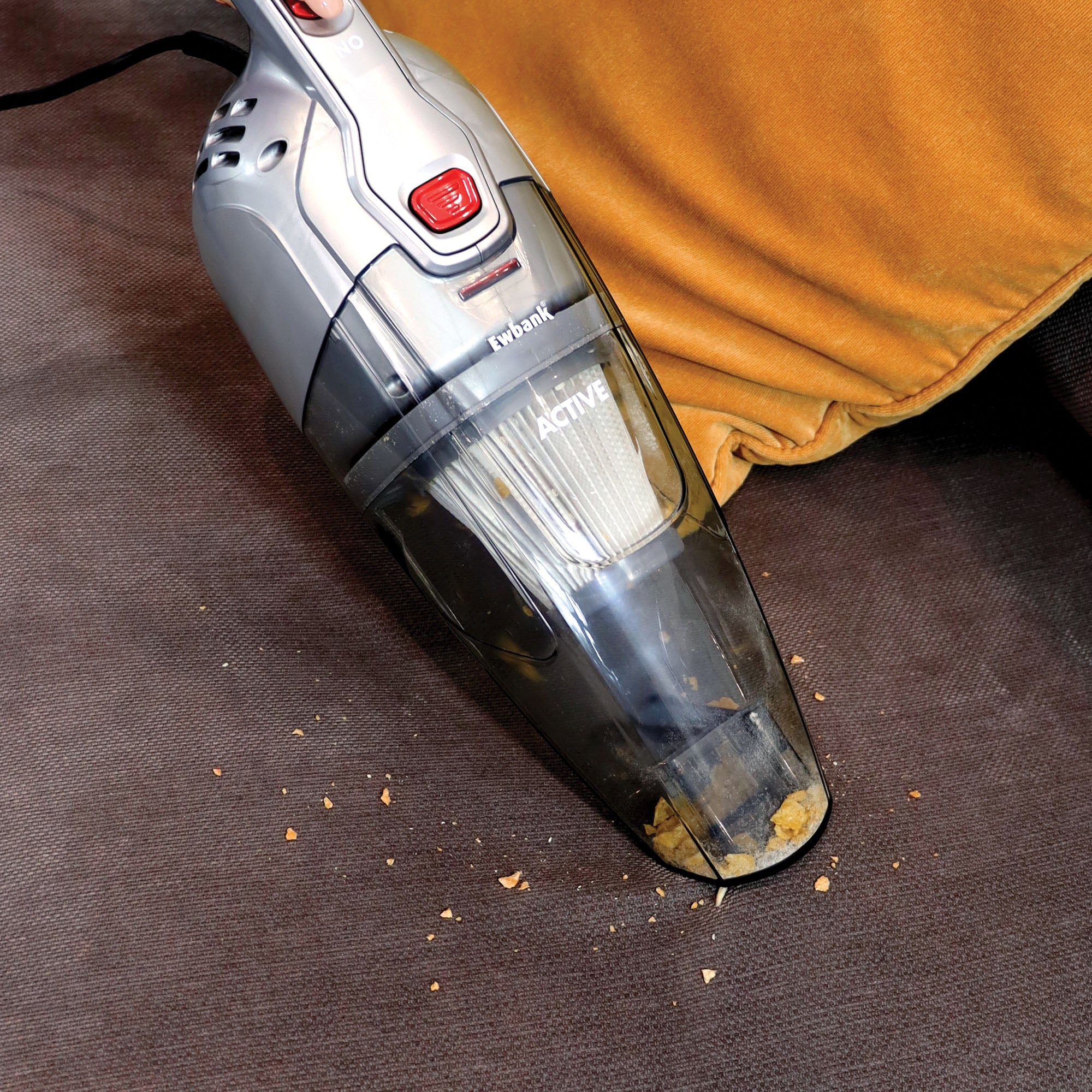 The ACTIVE 2-in-1 Corded Stick Vacuum Cleaner is used to clean crumbs from a brown fabric sofa beside a mustard yellow cushion, highlighting its versatility on various surfaces.