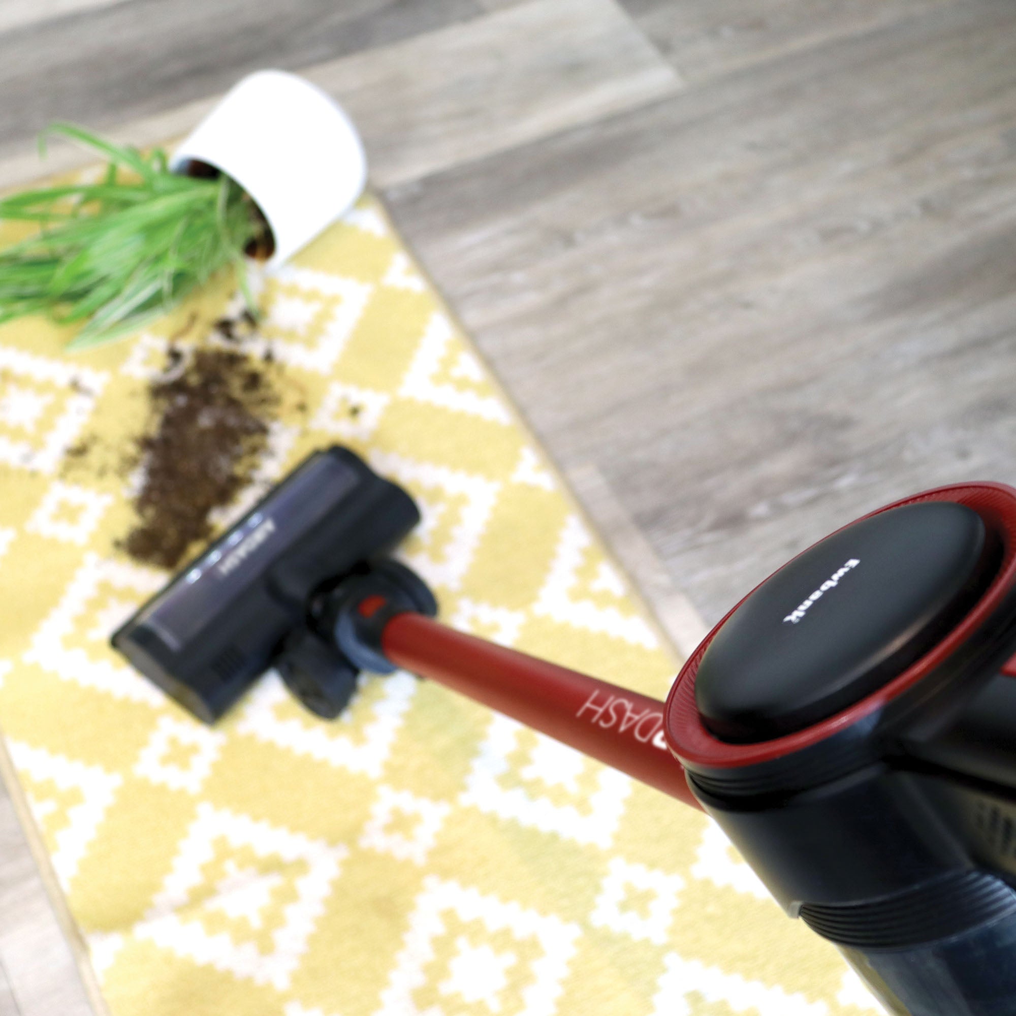 The AIRDASH 2-In-1 Cordless Stick Vacuum Cleaner is cleaning spilled soil from a fallen potted plant on a yellow patterned rug placed over wooden flooring.