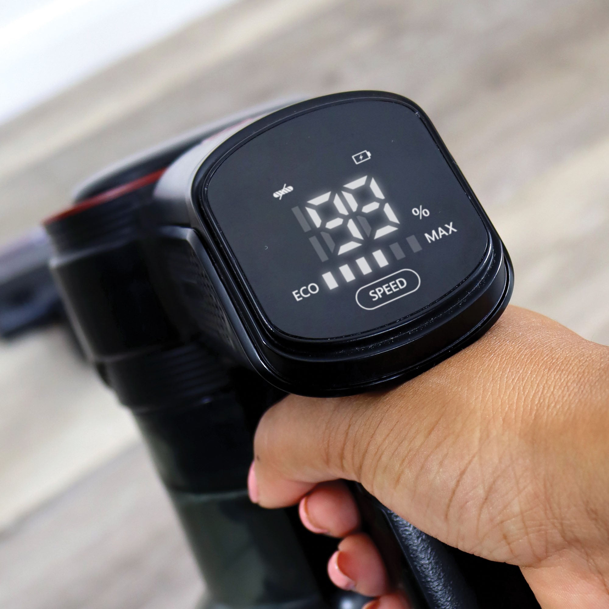 Close-up of a hand holding the AIRDASH 2-In-1 Cordless Stick Vacuum Cleaner, featuring a digital display with battery (100%) and speed indicators, eco and max modes, and a washable HEPA filter. The lightweight design stands out against a blurred background.