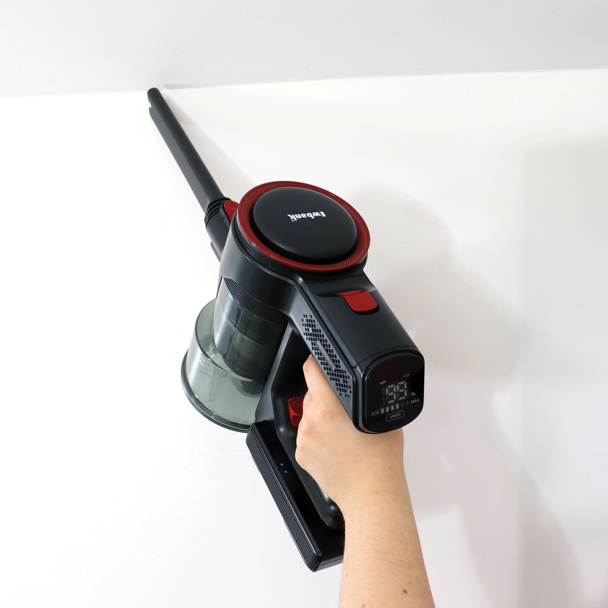 A person uses the AIRDASH 2-In-1 Cordless Stick Vacuum Cleaner with a long nozzle attachment to clean the upper corner of a white wall.