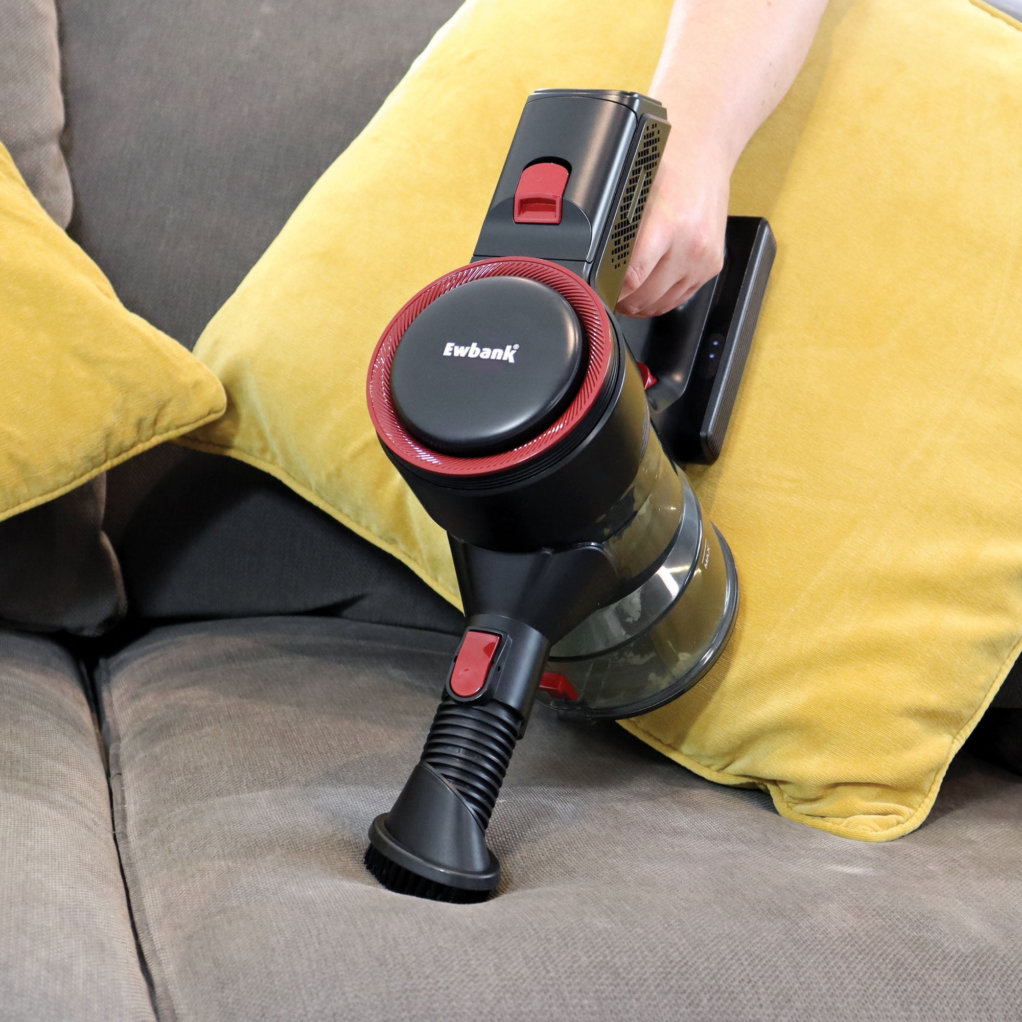 A person uses the AIRDASH 2-In-1 Cordless Stick Vacuum Cleaner with a brush attachment to clean a yellow cushion on a gray sofa.