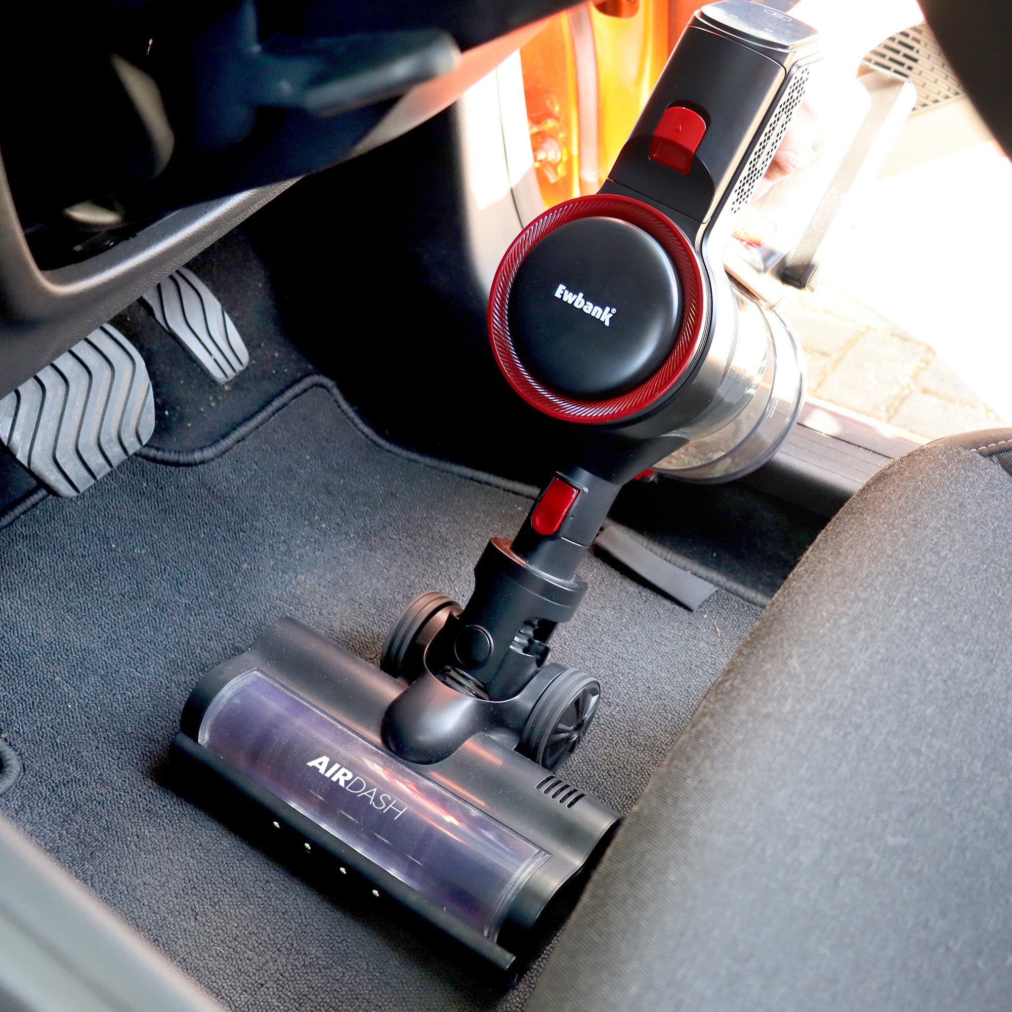 The AIRDASH 2-In-1 Cordless Stick Vacuum Cleaner is cleaning a car’s floor mat, with car pedals and part of the seat visible nearby.