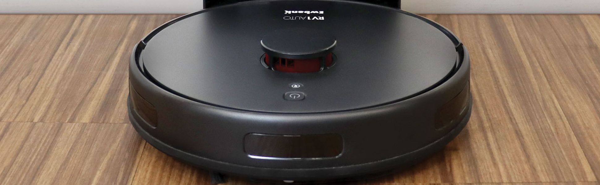 A black Ewbank RV1 Auto robot vacuum with an auto-empty station on a wooden floor, featuring icons and text highlighting its smart cleaning and 5000 PA suction power.