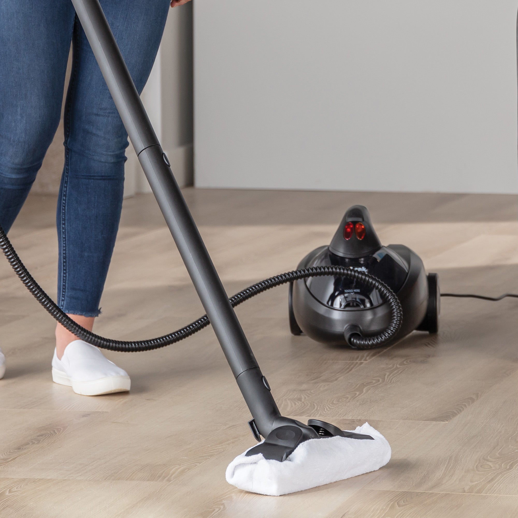 A person cleans light wood floors with only their lower body visible, using the STEAMDYNAMO+ 2000W Steam Cleaner & Sanitiser—its long-handled steam mop attaches by hose to the black, wheeled unit for chemical-free floor cleaning.