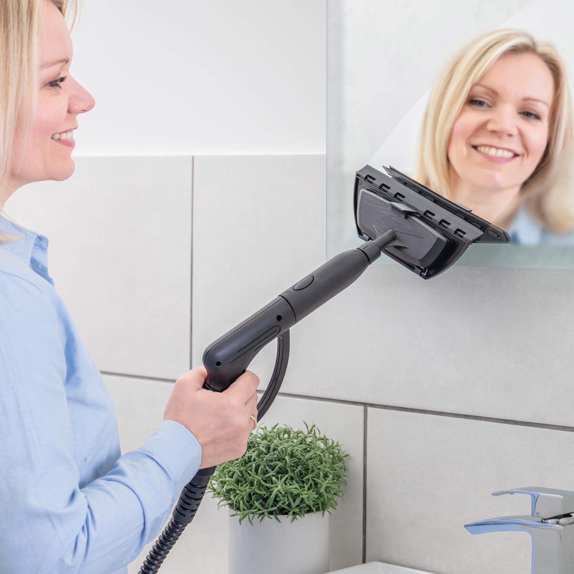 A woman smiles while cleaning a bathroom mirror with the STEAMDYNAMO+ 2000W Steam Cleaner & Sanitiser. On the sink below are a green potted plant, soap dispenser, and faucet, highlighting her commitment to chemical-free cleaning.