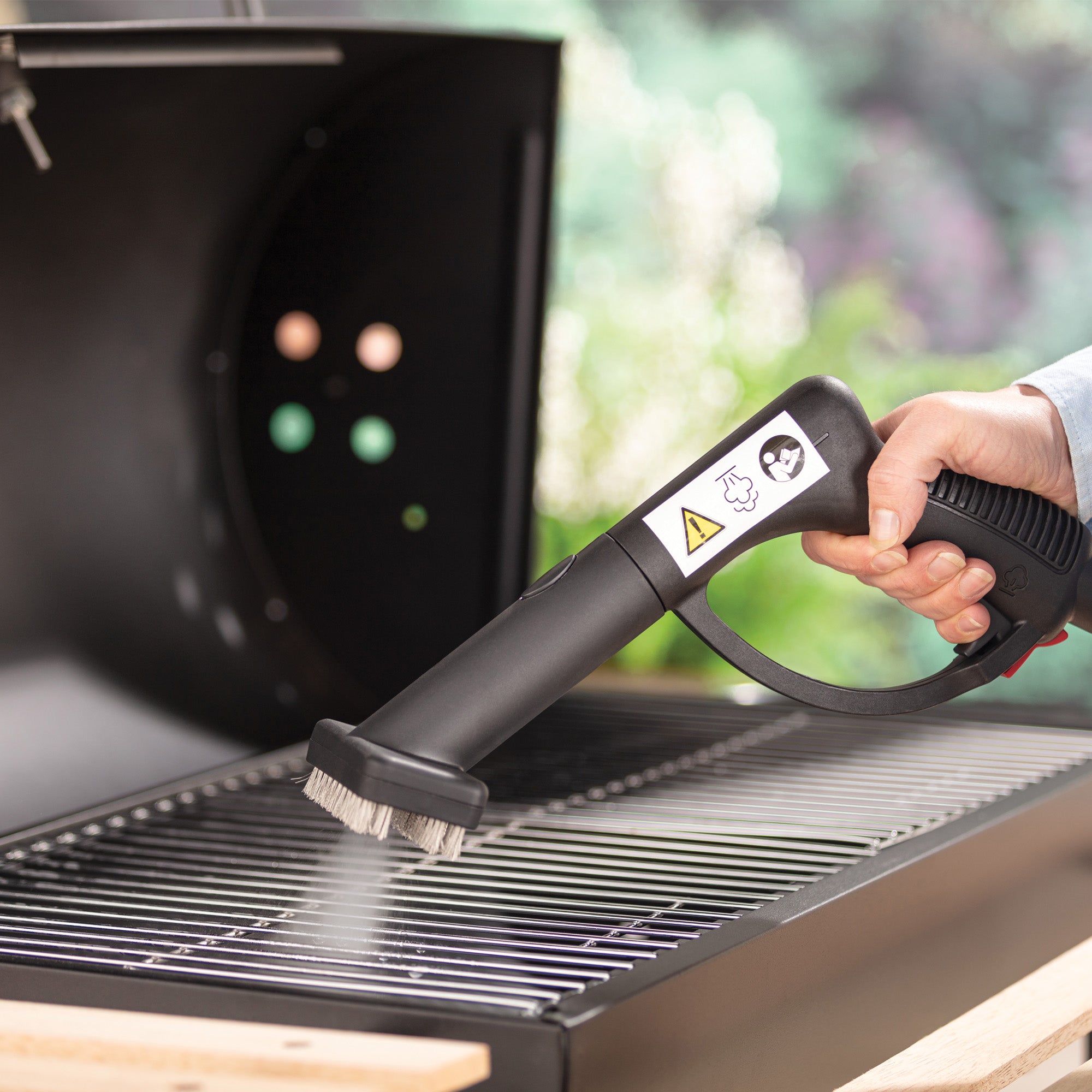 Using the STEAMDYNAMO+ 2000W Steam Cleaner & Sanitiser, a person cleans the metal grate of an outdoor barbecue grill with chemical-free steam. Steam is visible from the brush, and there's a warning label on the handle.