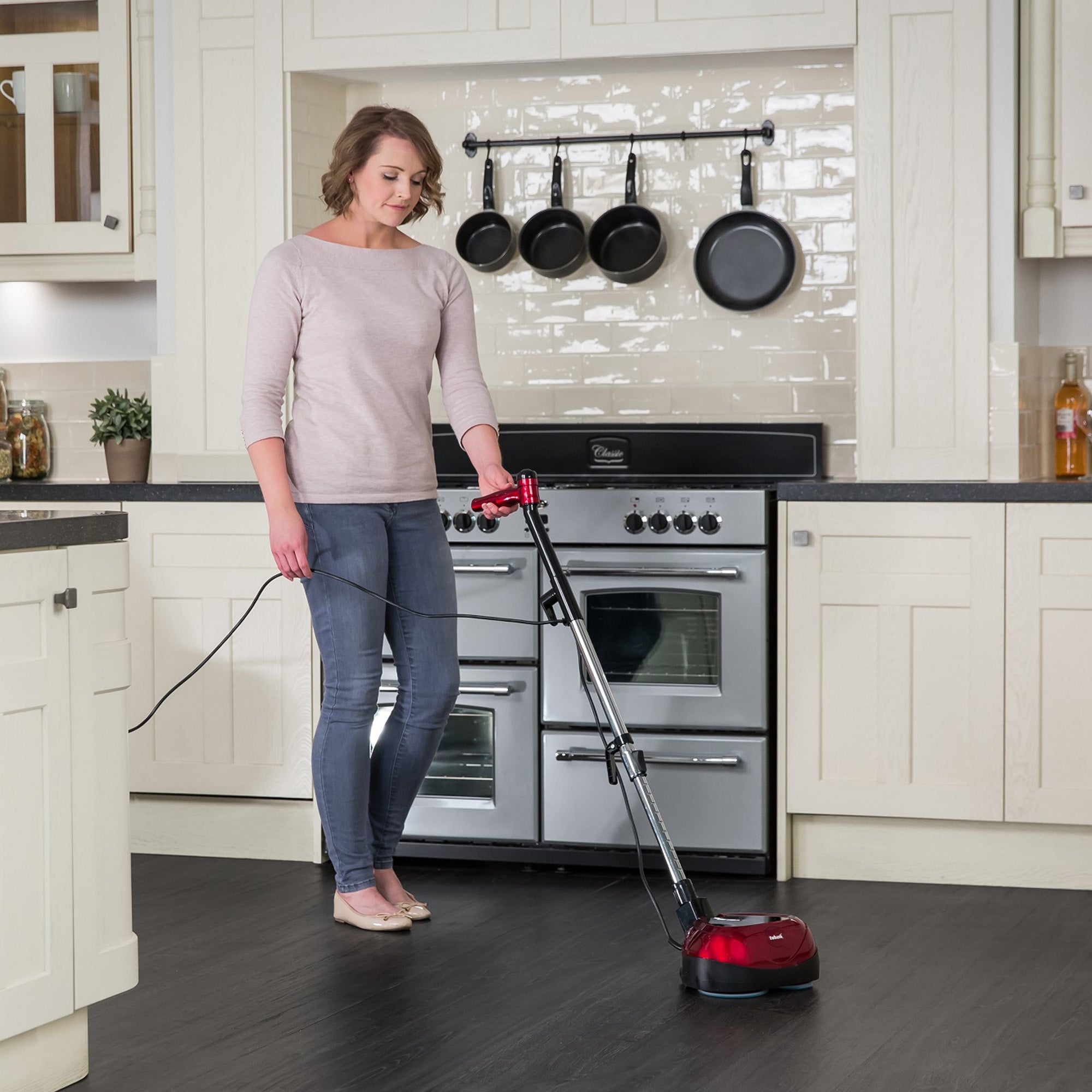 A woman in a light sweater and jeans uses the EP170 Multi Use 3-in-1 Floor Cleaner & Polisher on a dark kitchen floor, with white cabinets, a stove, and black pans hanging on a tiled wall behind her.
