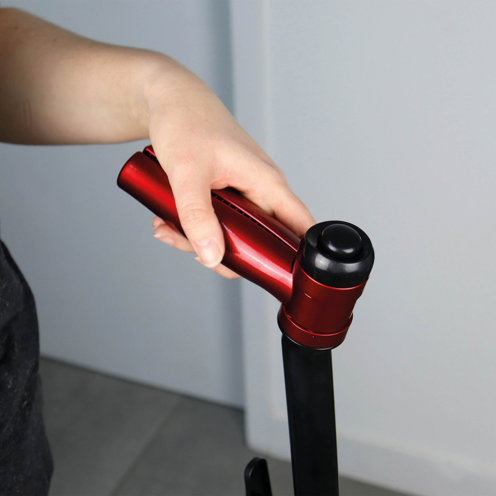 A person’s hand holds the red handle of a suitcase indoors near a white wall and gray floor tiles that gleam as if cleaned by the EP170 Multi Use 3-in-1 Floor Cleaner & Polisher.