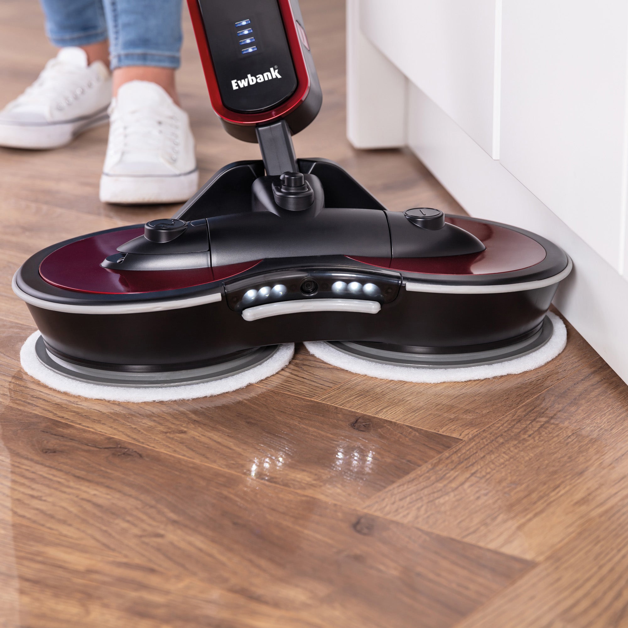 A person cleans a wood-patterned floor near a white kitchen cabinet edge using the FP90 Lightweight Cordless Floor Polisher & Cleaner, which features dual rotating pads, LED lights, and a rechargeable battery.