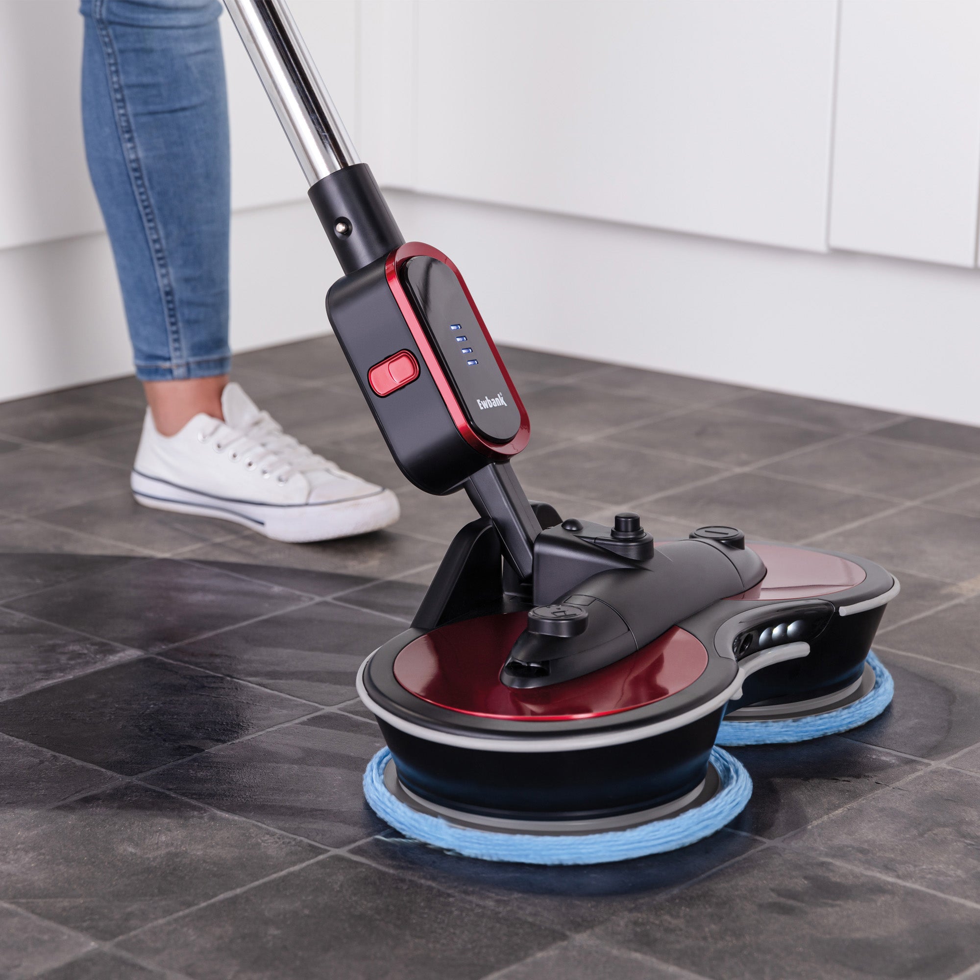 Wearing jeans and white sneakers, a person uses the FP90 Lightweight Cordless Floor Polisher & Cleaner with blue pads to clean a dark tiled floor in a modern kitchen.