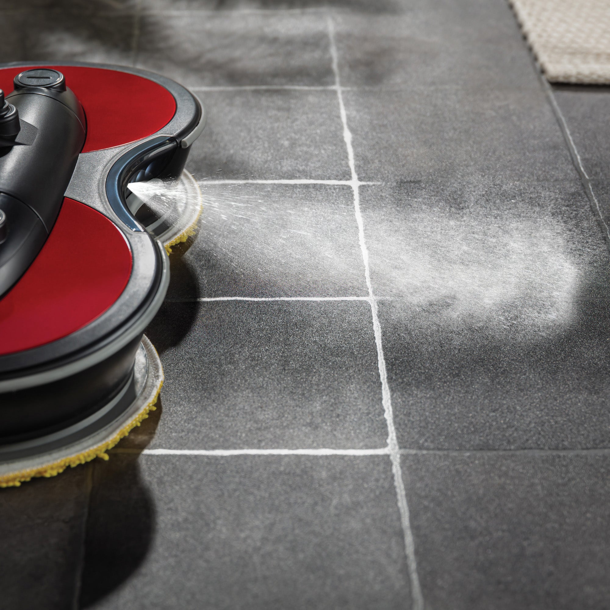 The FP90 Lightweight Cordless Floor Polisher & Cleaner, in red and black, sprays water as it moves across dark gray tiles, efficiently cleaning grout and surfaces with powerful commercial-level performance.