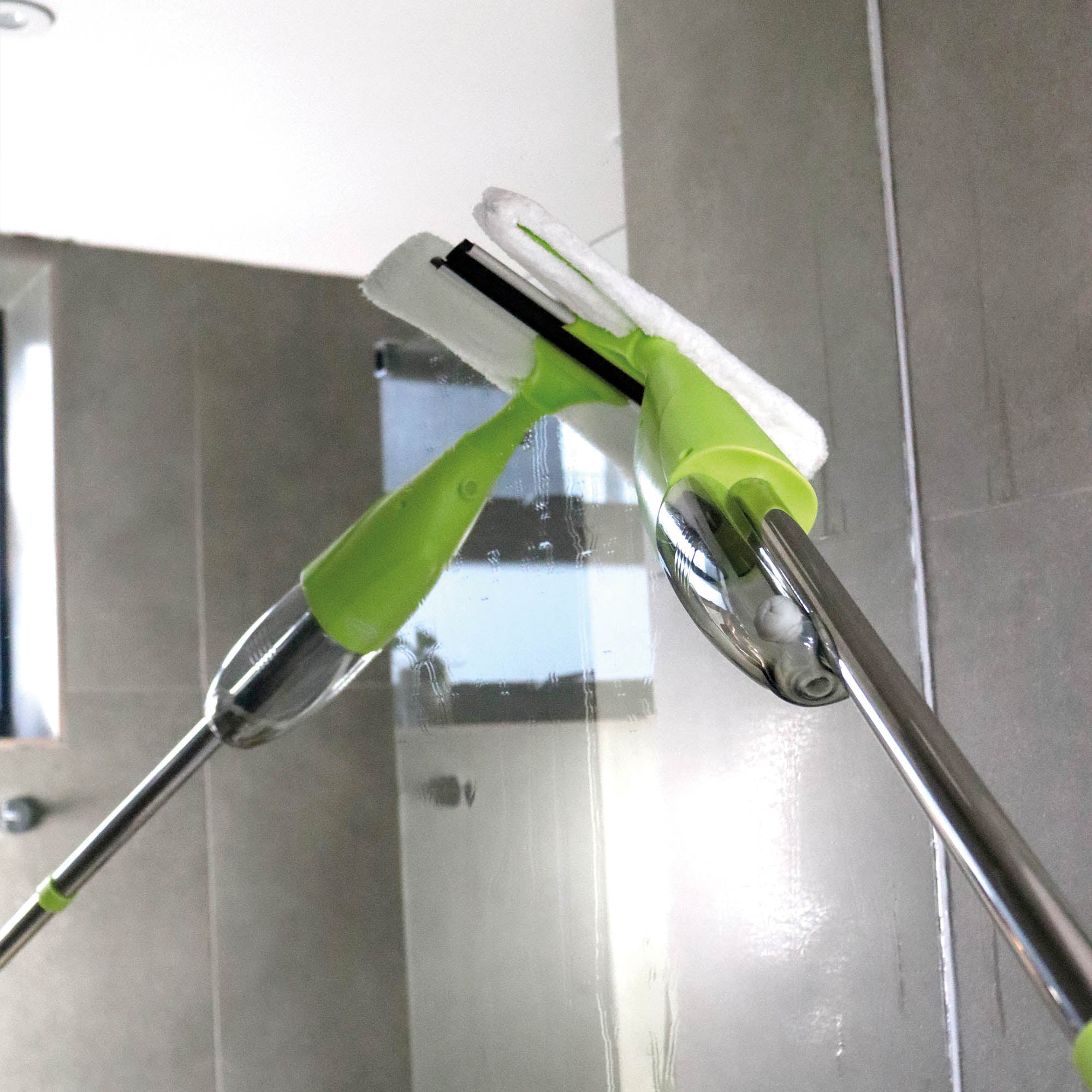 A close-up of the Ewbank 5-in-1 Universal Spray Mop Set in green with interchangeable heads, used to clean a glass shower door and remove streaks, with gray tiles in the background.