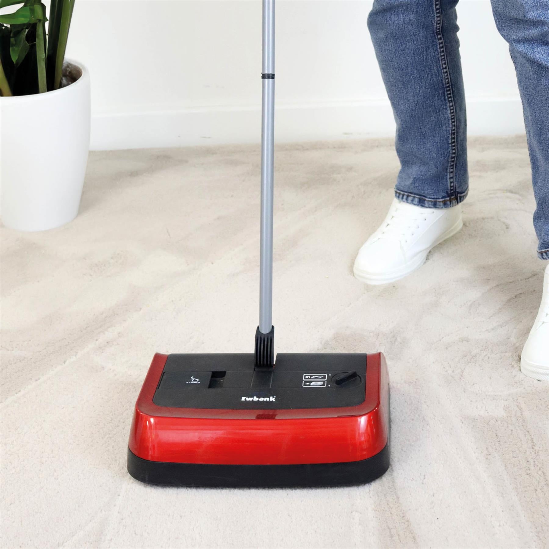 A person in blue jeans and white sneakers uses the red Evo3 Manual Carpet Sweeper with adjustable brush height on a light carpet, while a white plant pot is visible in the background.