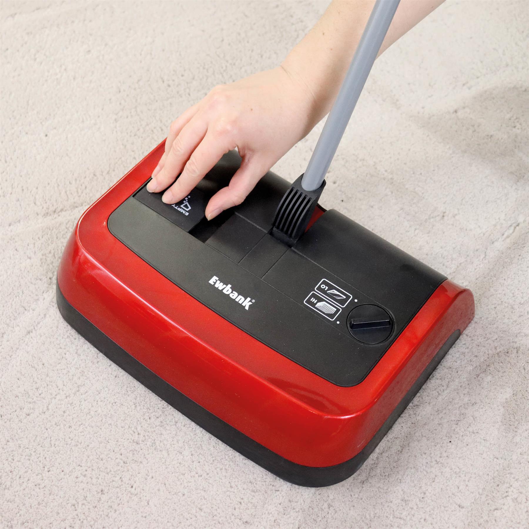 A hand adjusts the brush height on the red and black Evo3 Manual Carpet Sweeper with a gray handle, placed on a light beige carpet for improved hygienic cleaning.