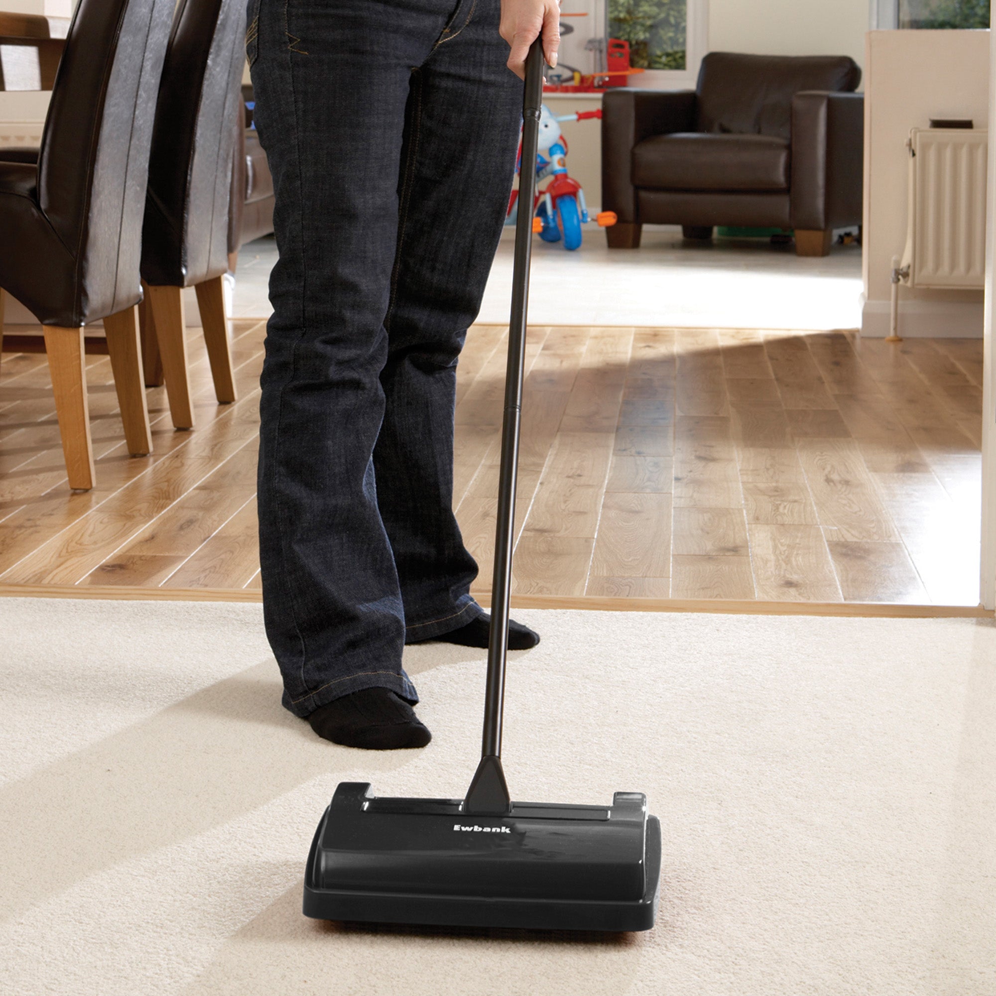 A person uses the SPEEDSWEEP Manual Carpet Sweeper by Ewbank (black, adjustable height) on a light-colored carpet in a living room with wooden floors and furniture visible in the background.