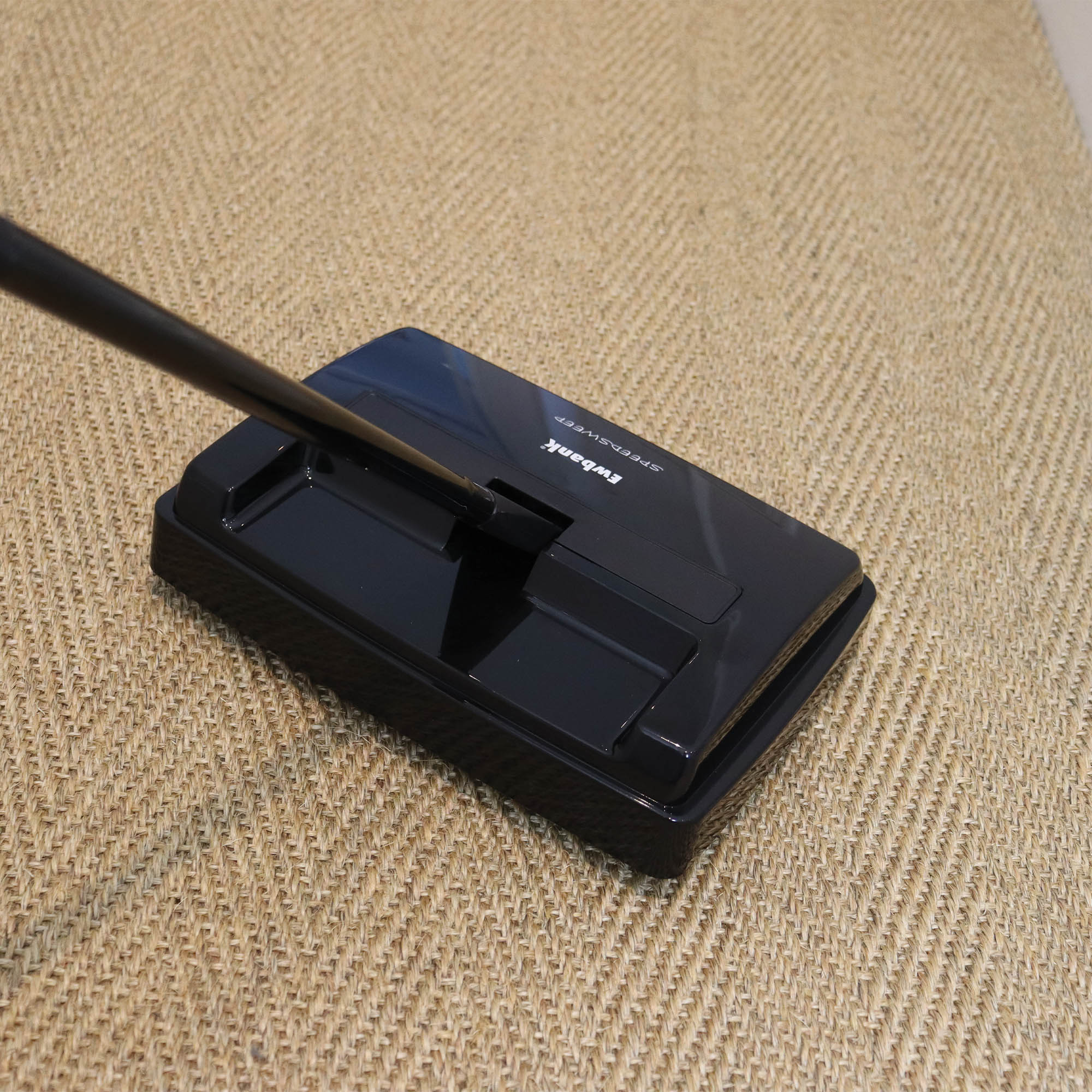 The SPEEDSWEEP Manual Carpet Sweeper by Ewbank, featuring adjustable height and a compact black design, is being used on a beige textured carpet.
