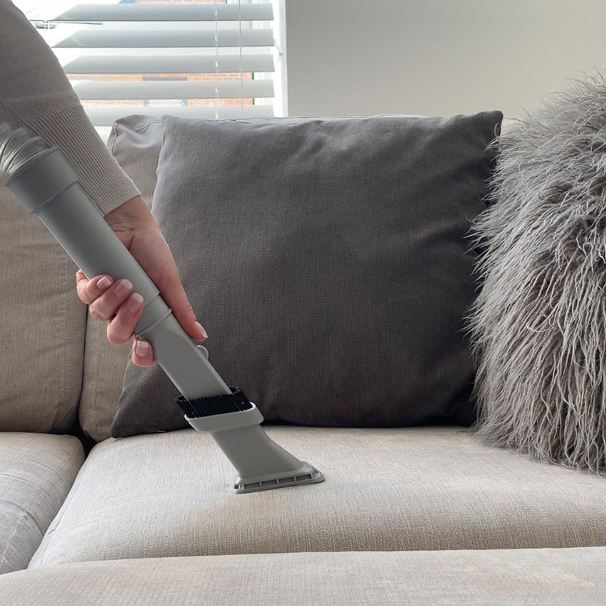 Using the MOTION Pet Upright Bagless Vacuum Cleaner with a turbo brush, a person cleans a light-colored fabric sofa decorated with gray fluffy pillows in a bright living room.