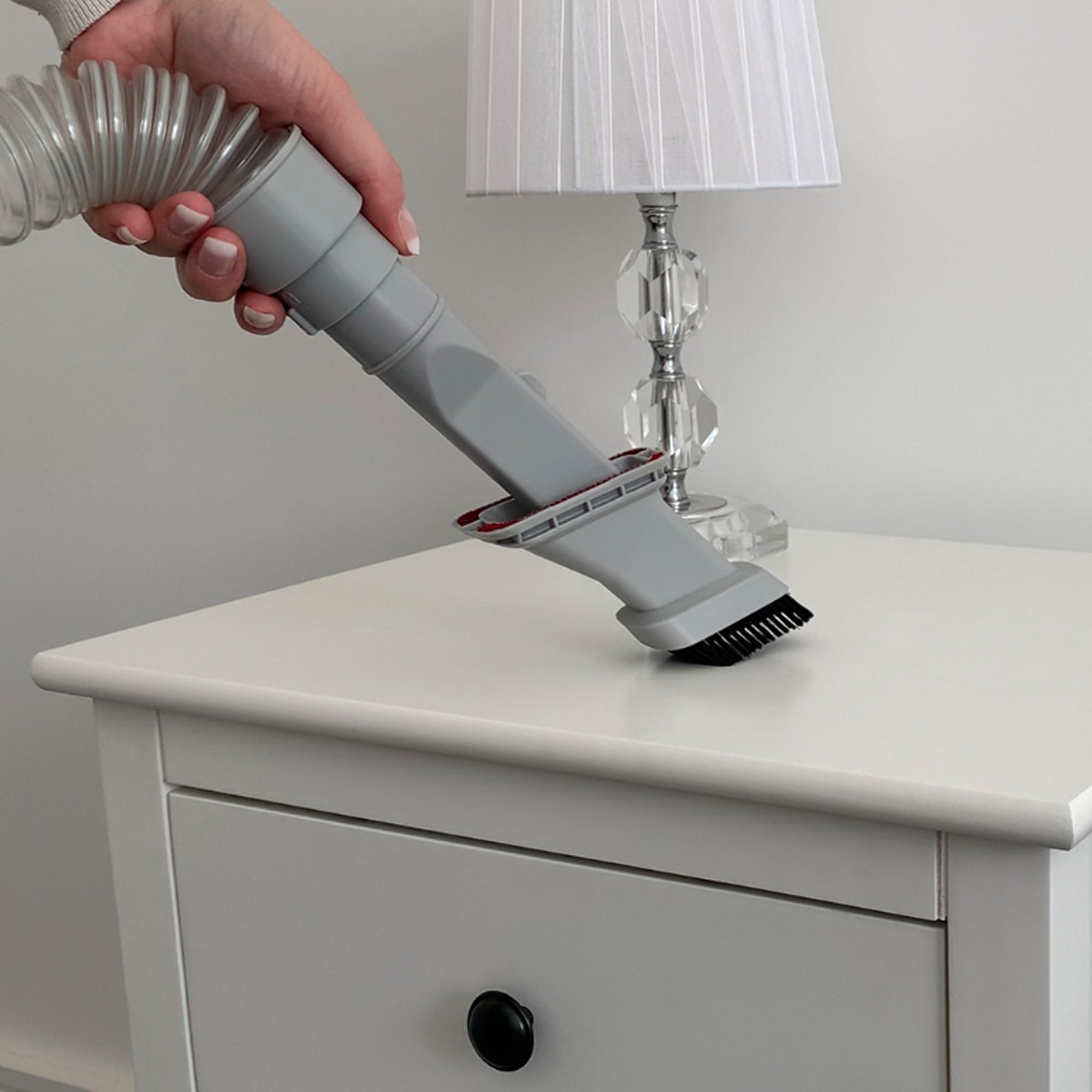 A hand uses the MOTION Pet Upright Bagless Vacuum Cleaner with a turbo brush to clean a white nightstand with a black knob; on top is a clear glass lamp with a white shade.