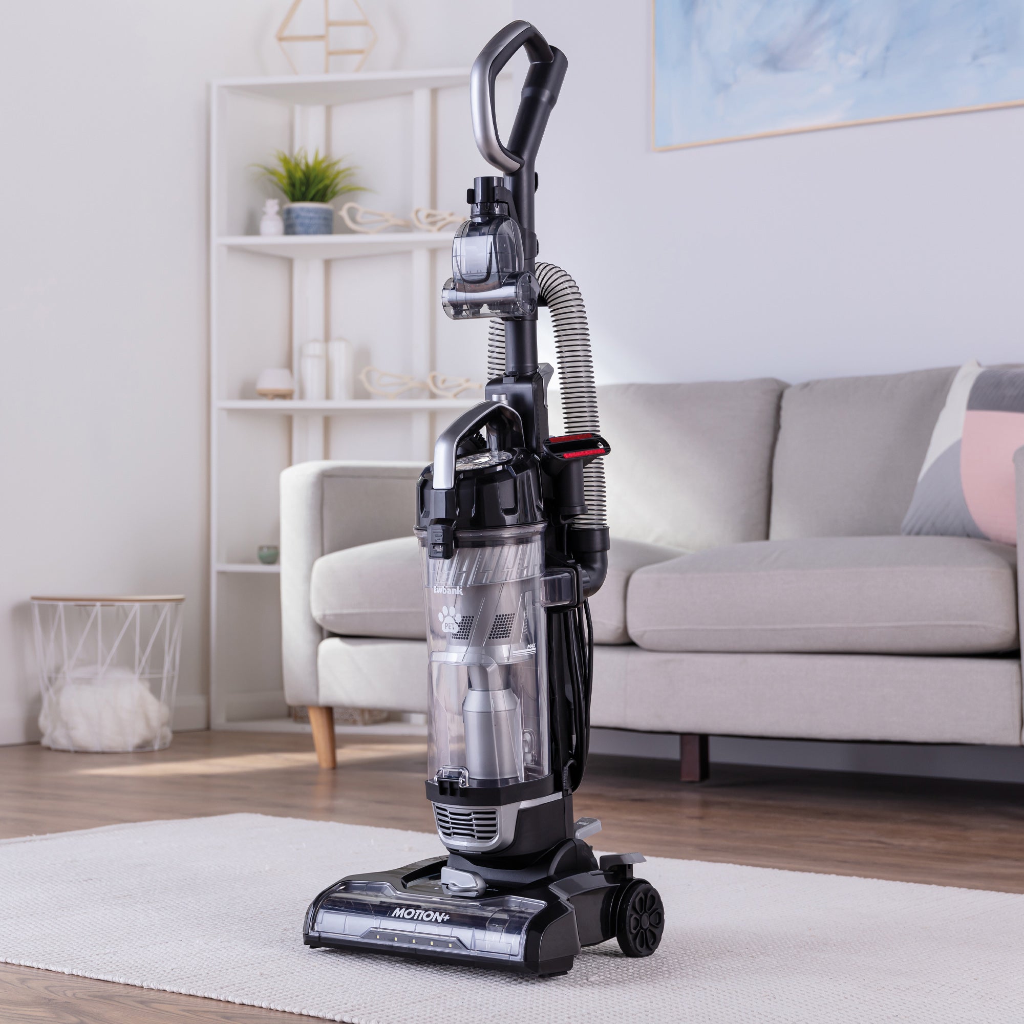 The MOTION+ Reach Pet Upright Bagless Vacuum Cleaner in black and gray stands ready on a wooden floor in a modern living room beside a light gray sofa, shelves, and elegant decor accents.
