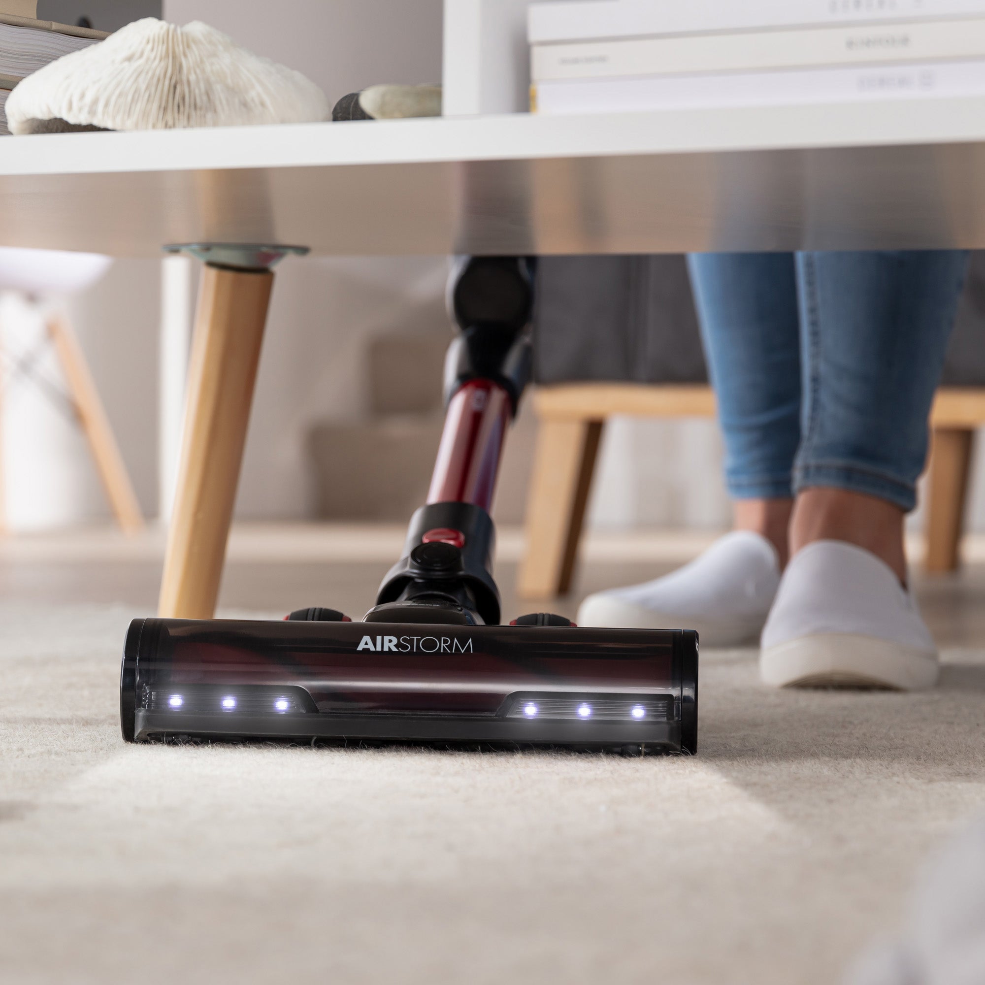 Wearing white slippers, a person uses the AIRSTORM 2-in-1 Cordless Stick Vacuum Cleaner with LED lights and a brushless DC motor to clean under a white coffee table on a carpeted floor.