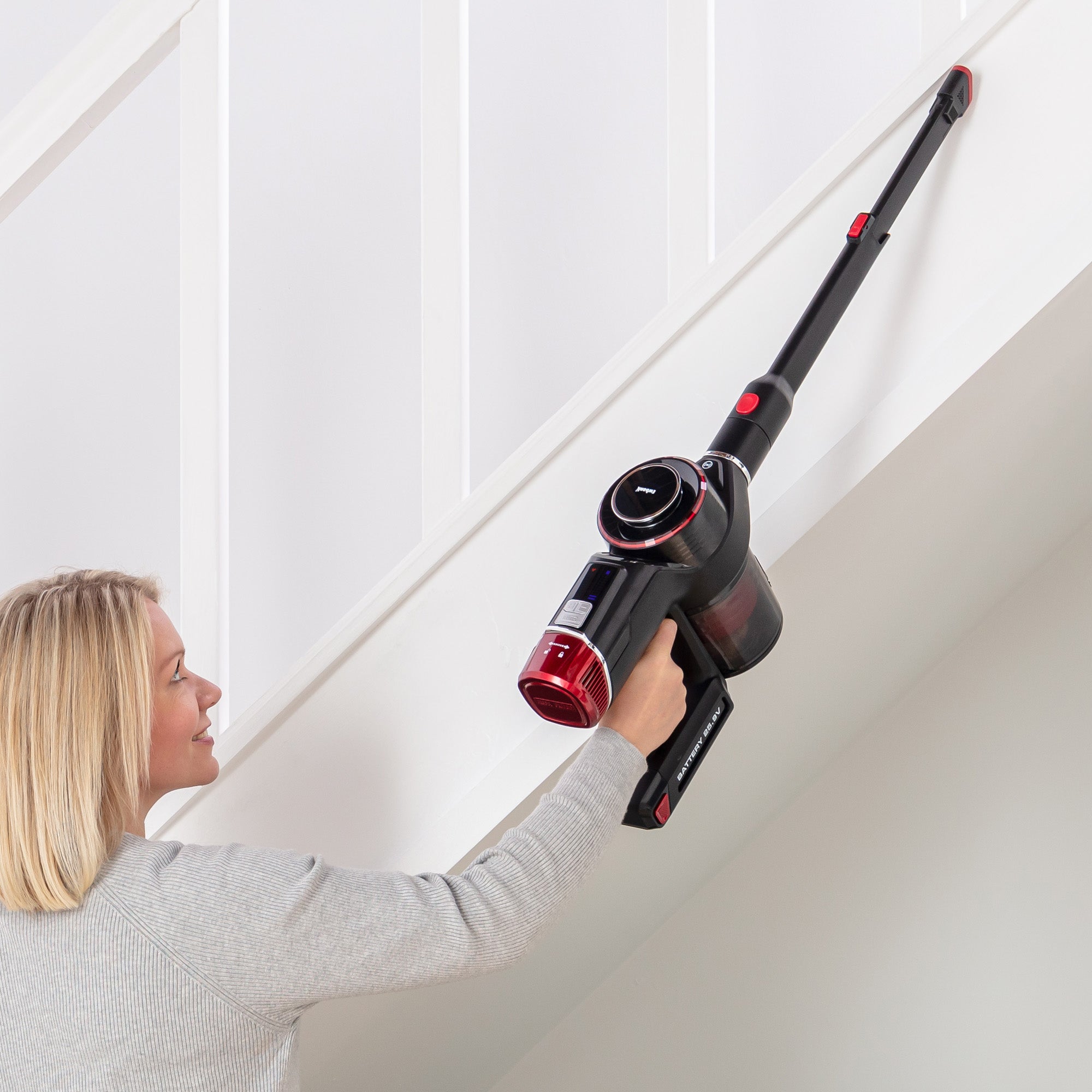 A woman uses the AIRSTORM 2-in-1 Cordless Stick Vacuum Cleaner, which has a red and black design and a brushless DC motor, to clean the white underside of a staircase.