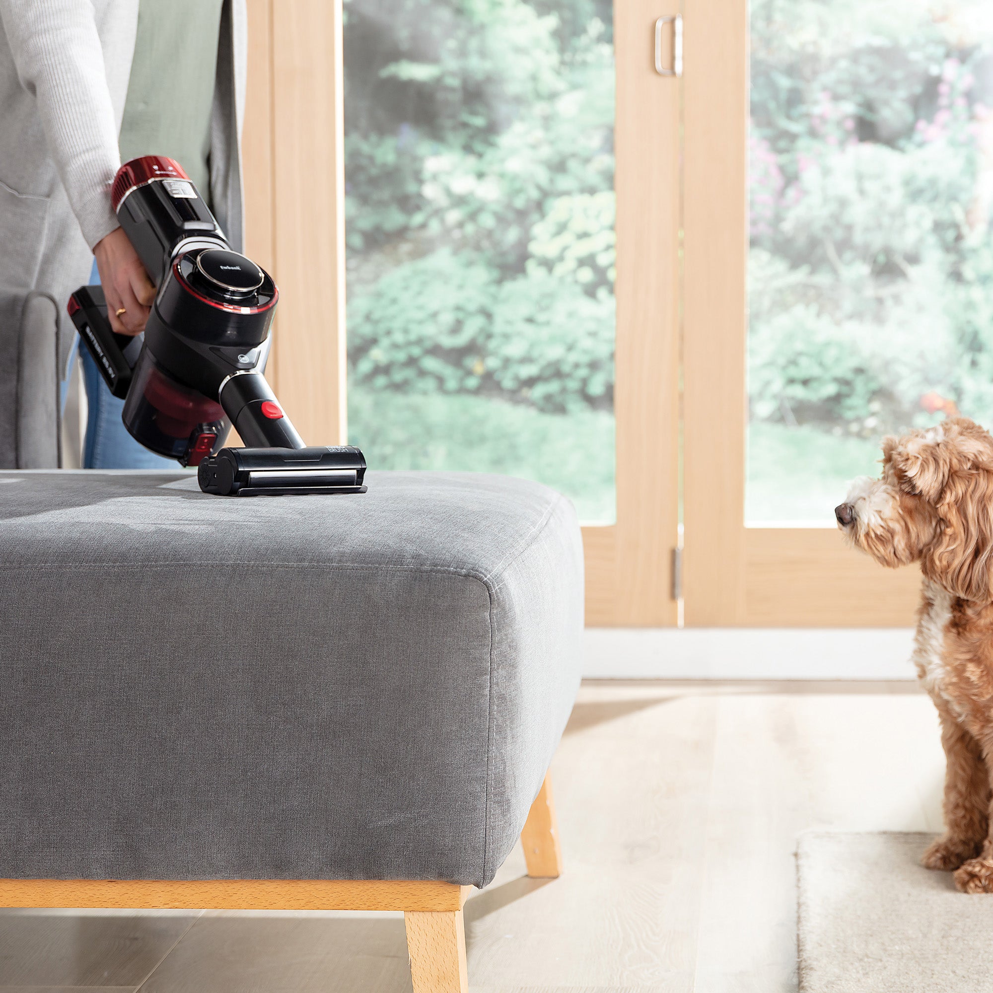 A person uses the AIRSTORM 2-in-1 Cordless Stick Vacuum Cleaner with a brushless DC motor on a gray ottoman, while a small brown and white dog sits on a light rug nearby. Large windows reveal a garden view in the background.