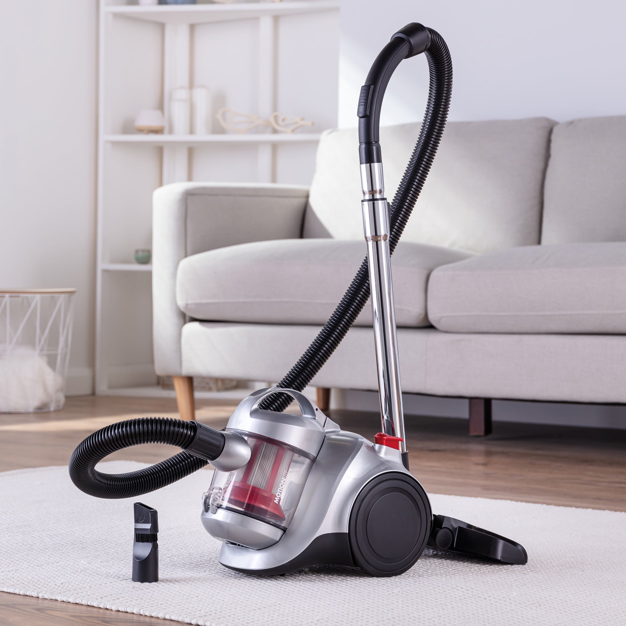The Ewbank MOTIONLITE Bagless Cylinder Vacuum Cleaner, featuring a hose and attachments, is placed on a light-colored rug in a living room with a gray sofa and shelves in the background.