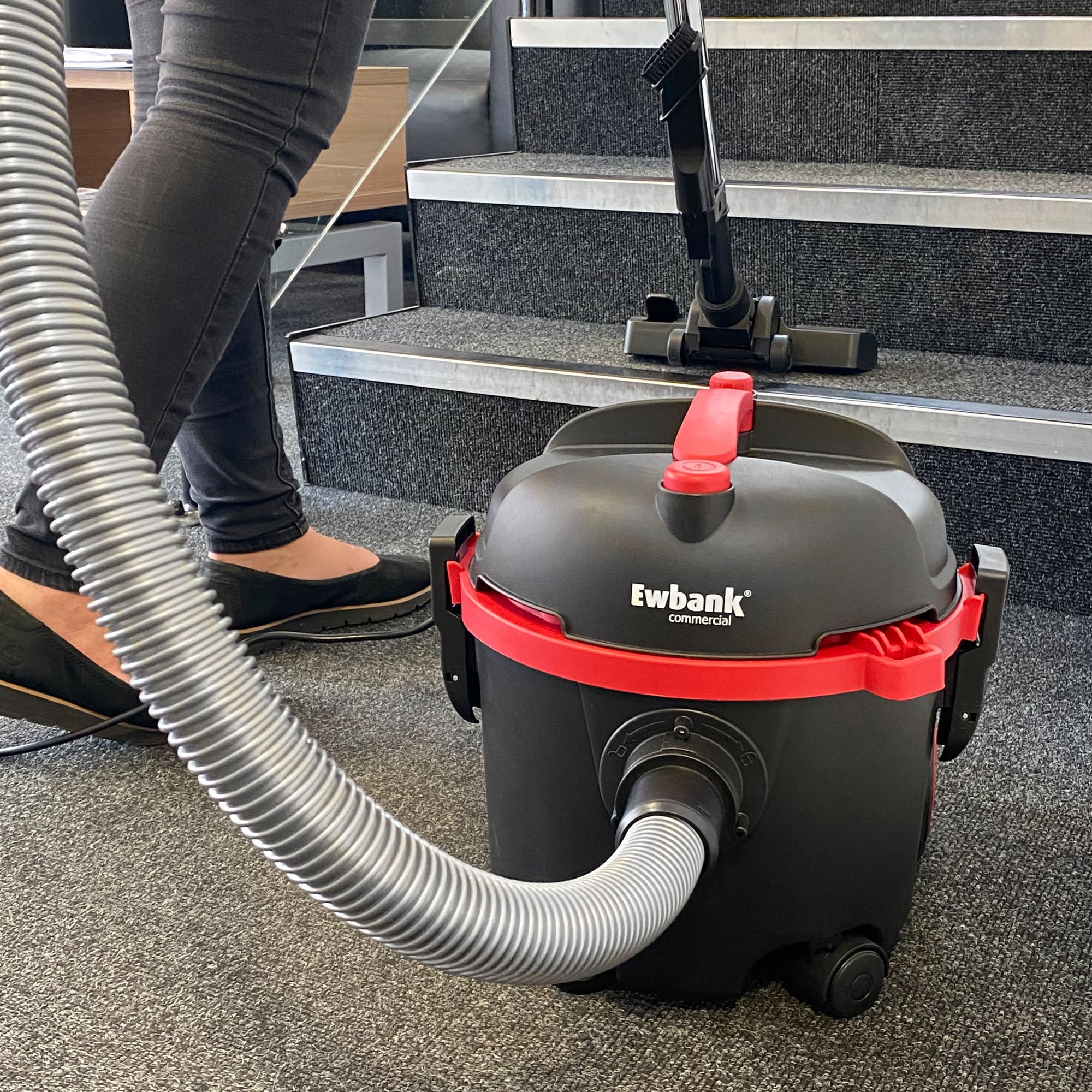 A person uses a black and red DV6 Dry Drum Vacuum Cleaner with a high-efficiency floor brush to vacuum carpeted stairs; the hose is extended, showing only their legs and feet.