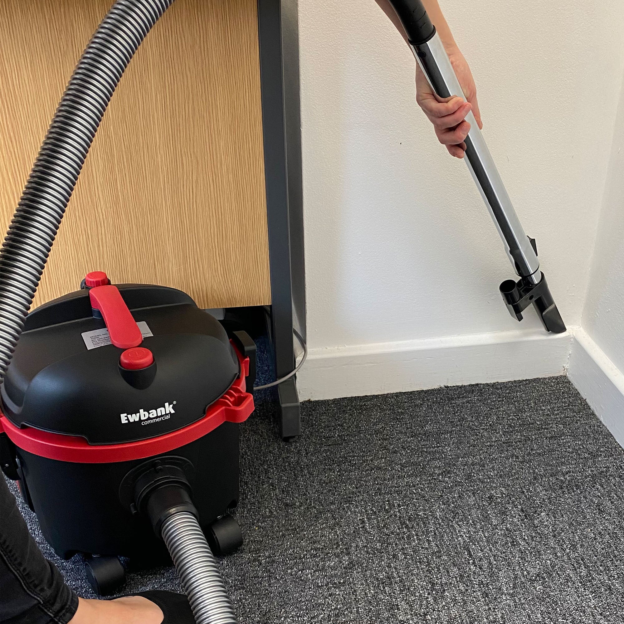 A person uses the DV6 Dry Drum Vacuum Cleaner with a high-efficiency floor brush and hose to clean the carpeted corner near a wooden desk and white wall.