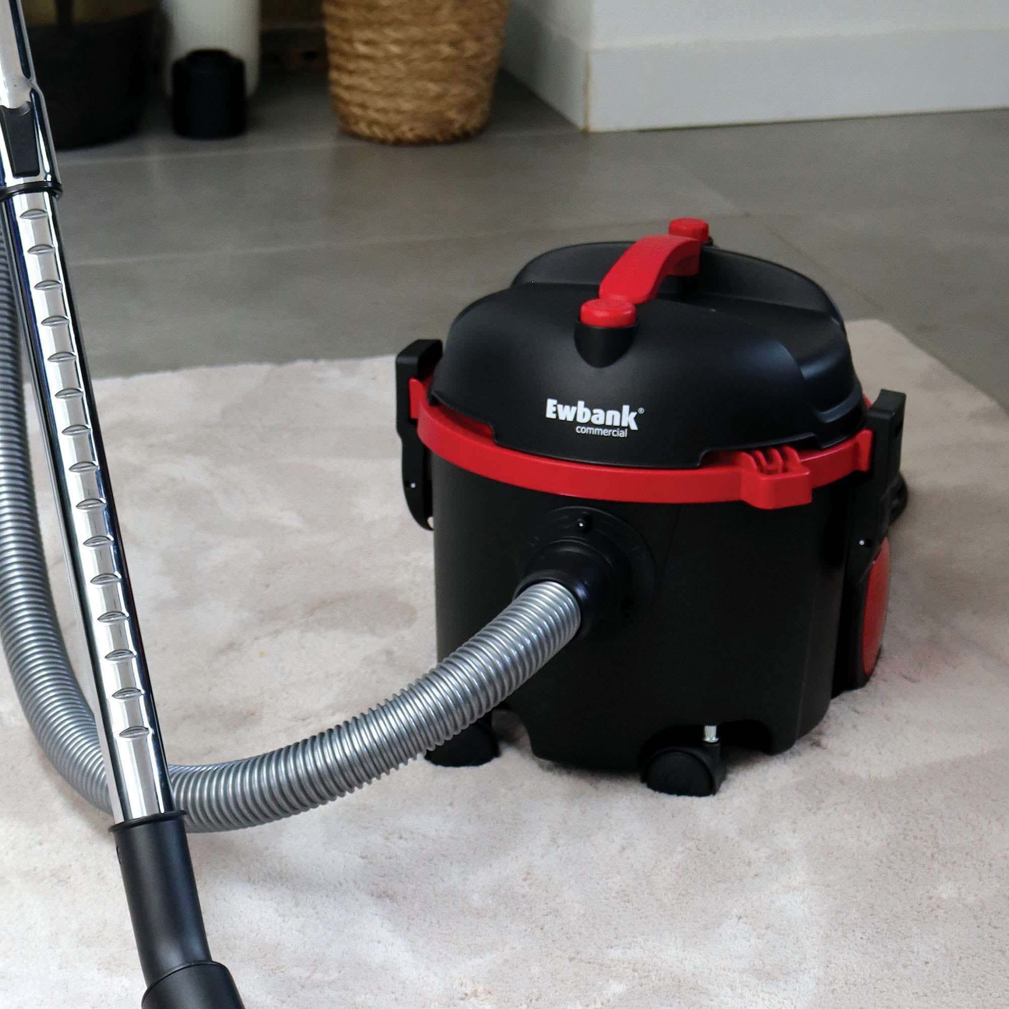 The black and red Ewbank DV6 Dry Drum Vacuum Cleaner with a high-efficiency floor brush sits on a light-colored rug, its hose and handle visible in the foreground next to a basket and cleaning supplies in the background.