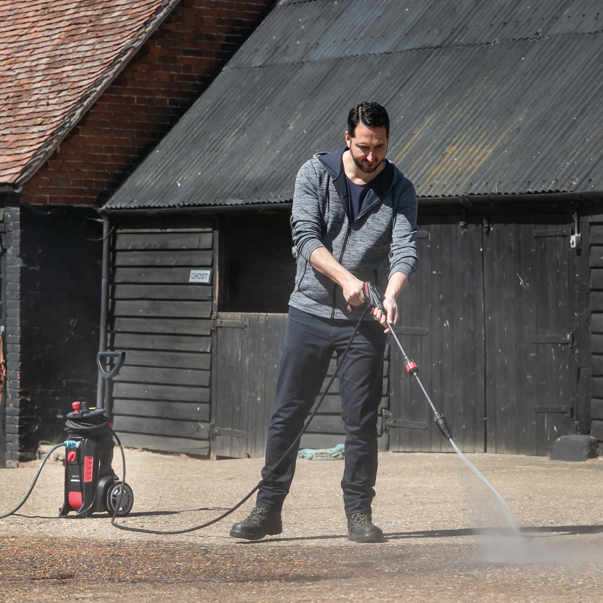 A man in dark pants and a gray hoodie uses the AQUABLAST160 High Power Pressure Washer (160/2320 PSI) to clean an outdoor surface in front of a dark wooden building.