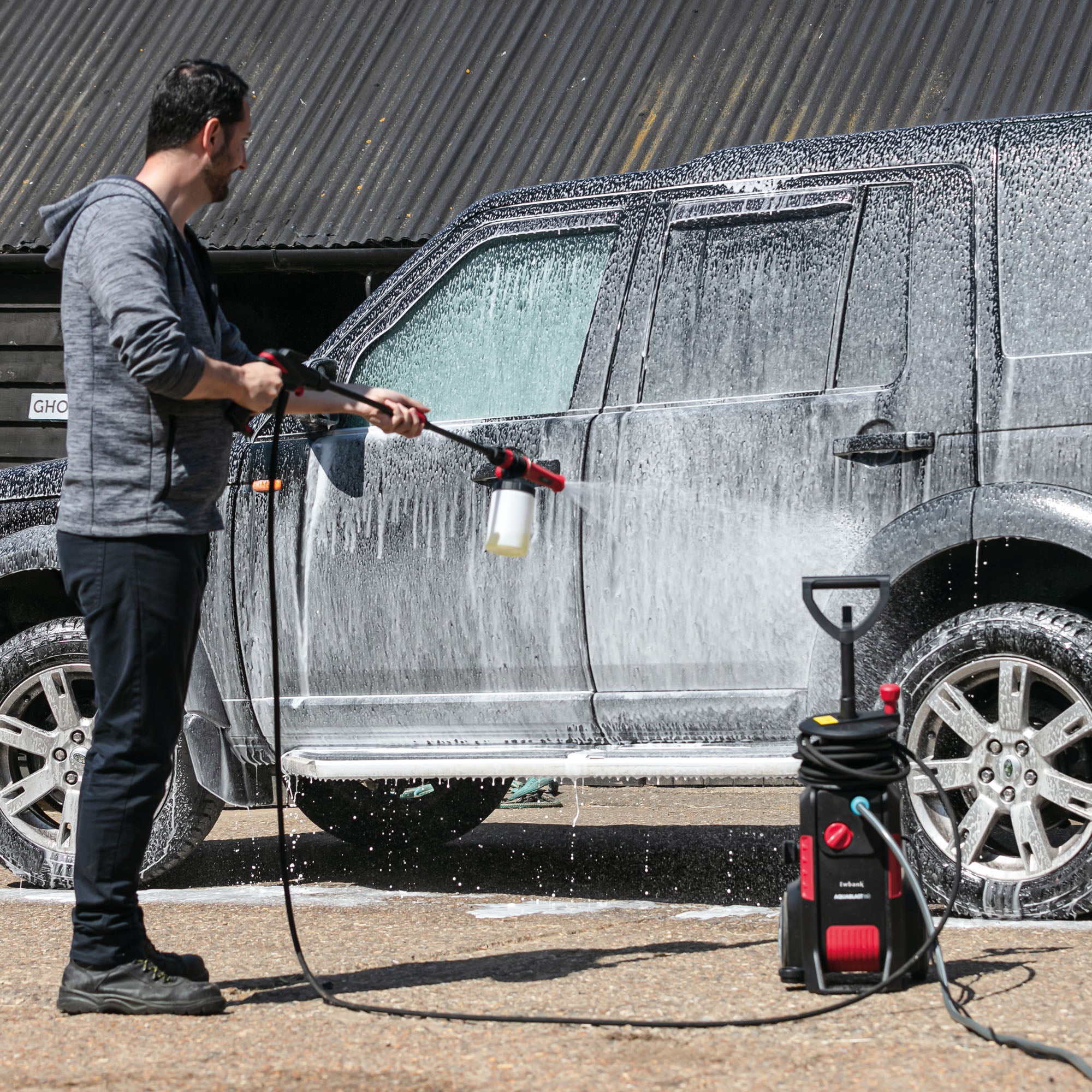 A man uses the AQUABLAST160 High Power Pressure Washer (160/2320 PSI) with a snow foam attachment to coat a black SUV in thick white suds, cleaning the car outdoors with equipment visible on the ground nearby.