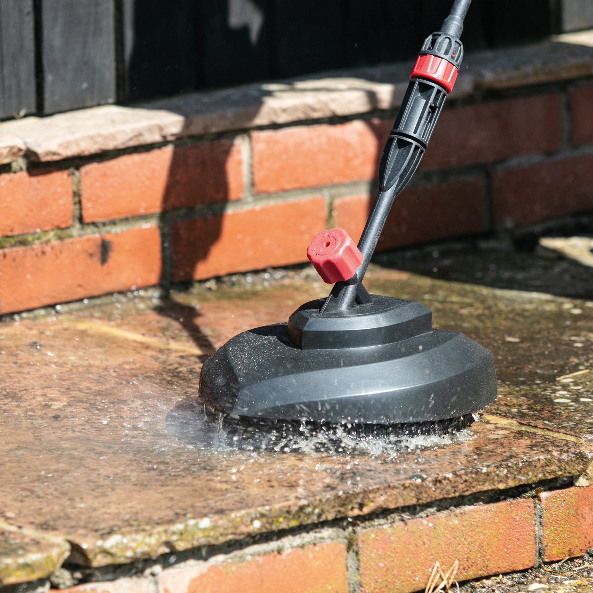 The AQUABLAST160 High Power Pressure Washer (160/2320 PSI) with a round surface cleaner blasts dirt and debris from a stone patio near a brick wall, spraying water around the cleaner during the cleaning process.