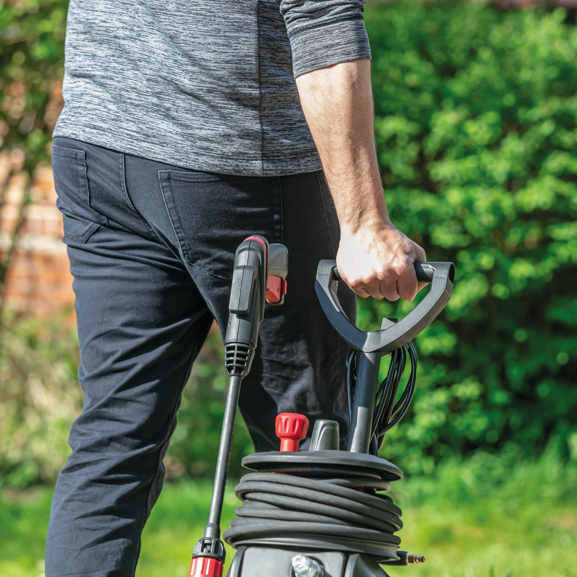 A person in dark pants and a gray shirt pulls the AQUABLAST160 High Power Pressure Washer (160/2320 PSI) with coiled hoses and red nozzles outdoors, green foliage in the background.