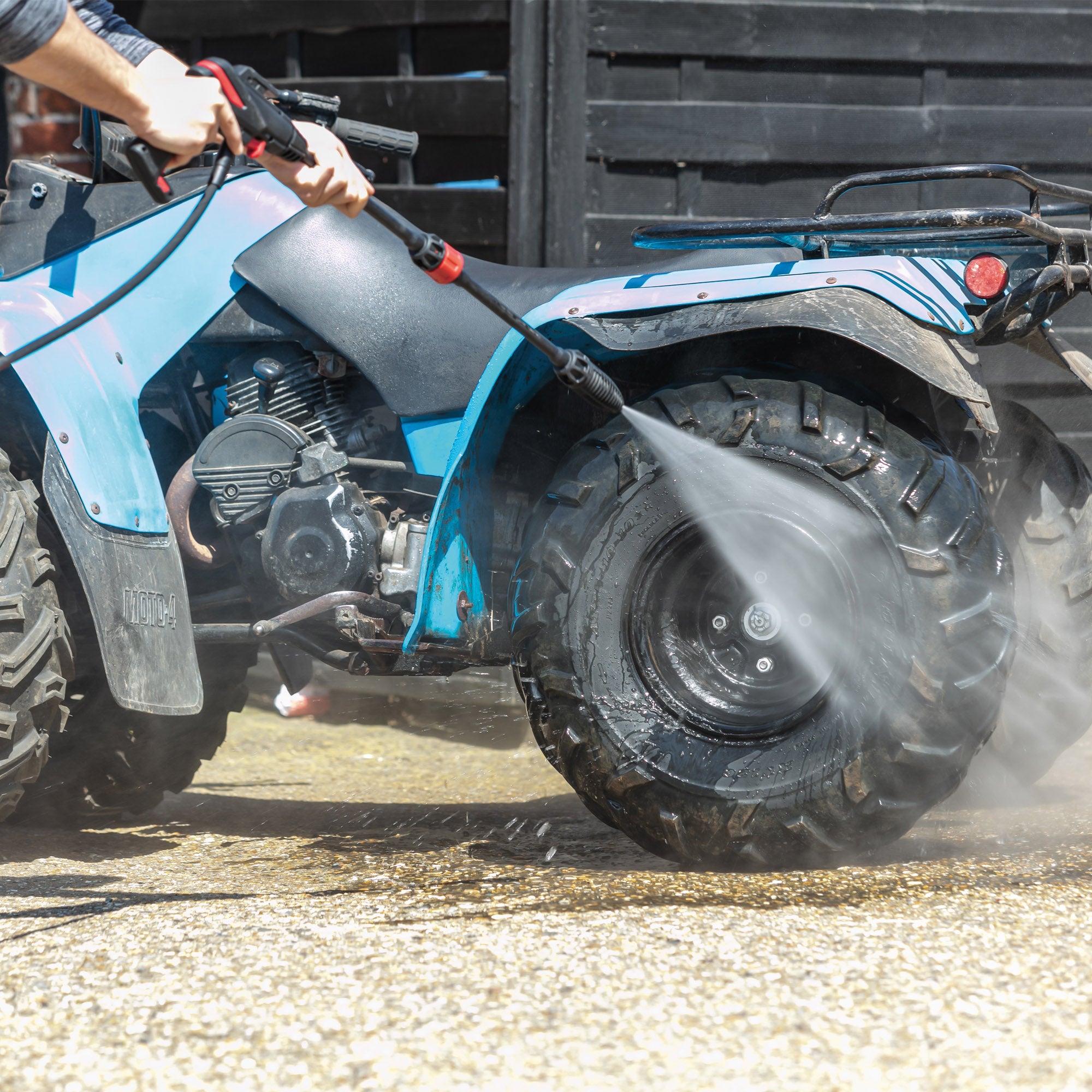 Using the AQUABLAST160 High Power Pressure Washer, 160/2320 PSI, a person cleans the large, muddy rear tire of a blue ATV outdoors on a sunny day.