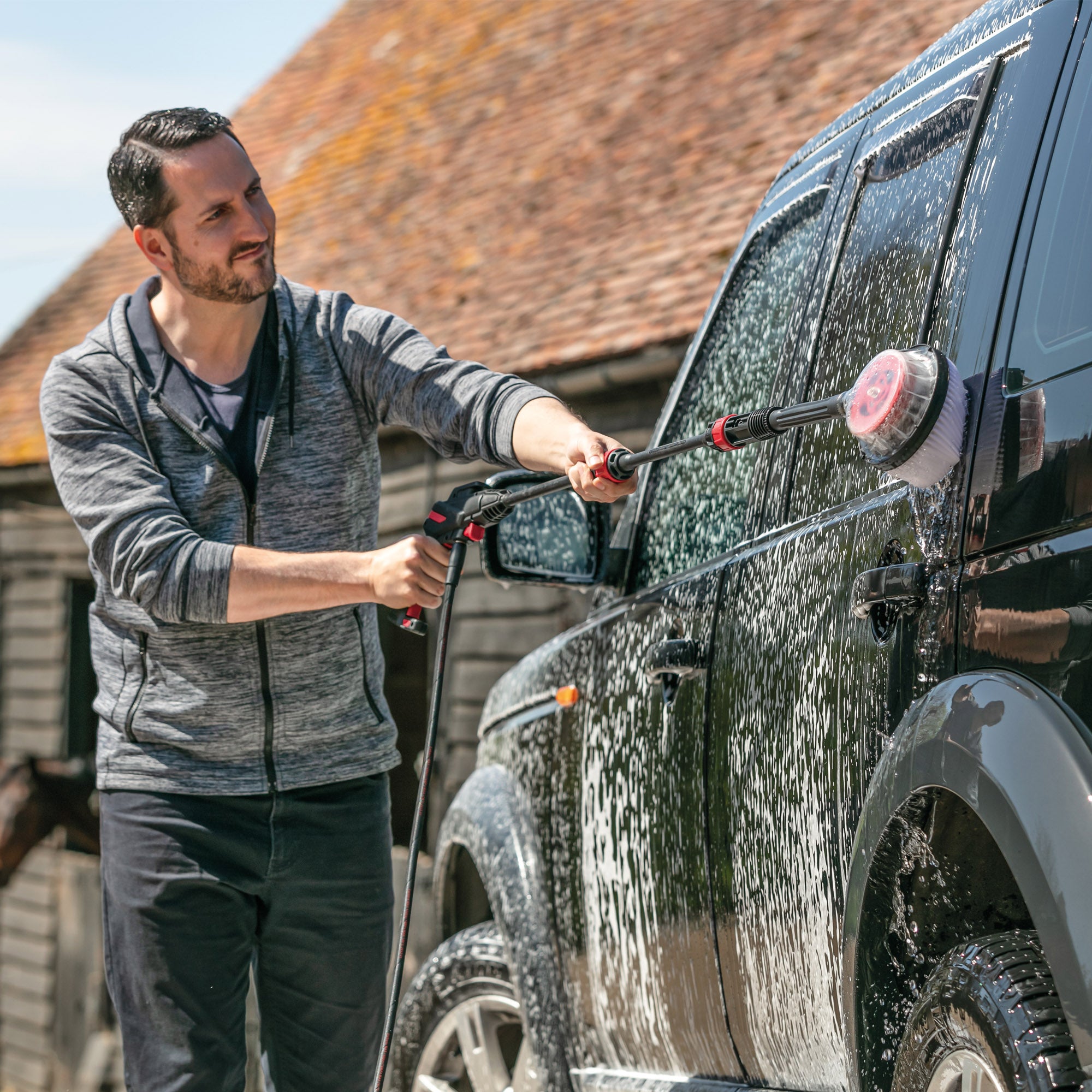 A man in a gray hoodie uses the AQUABLAST160 High Power Pressure Washer (160/2320 PSI) with a brush attachment to clean a soap-covered black SUV outdoors on a sunny day.