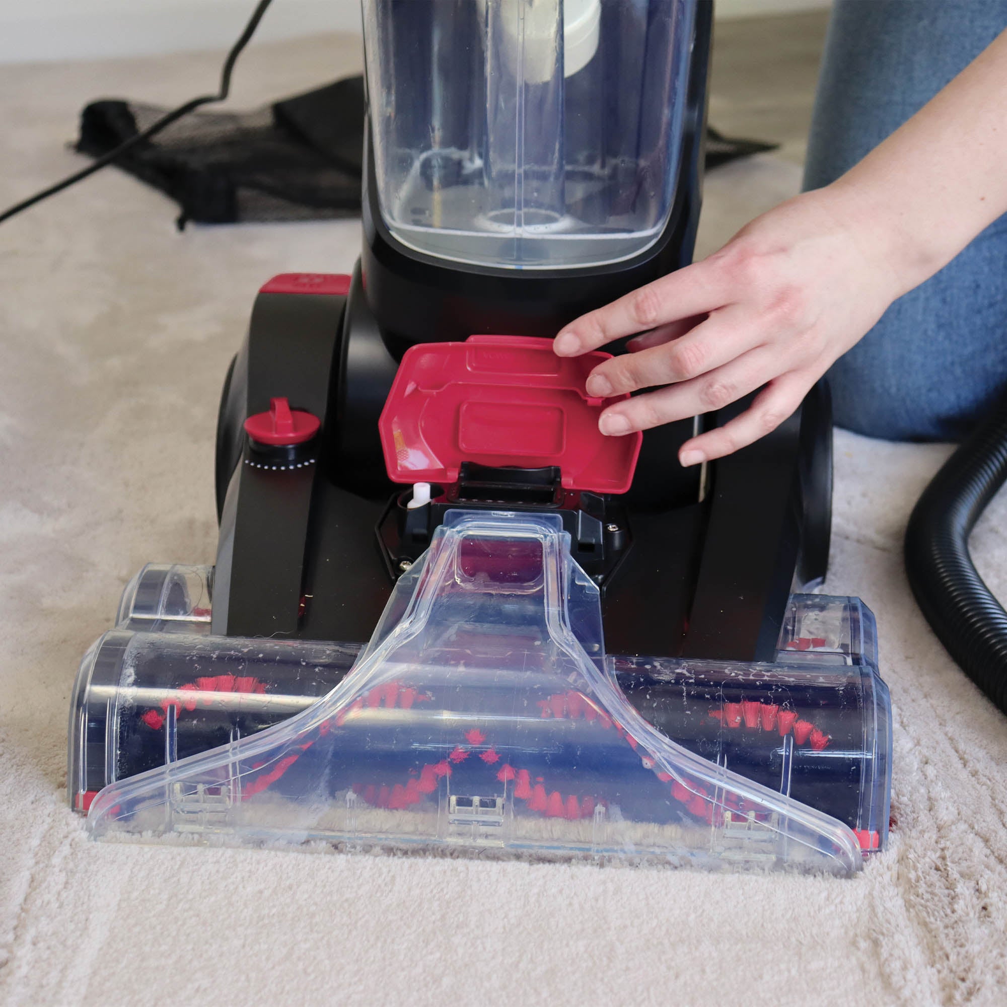 A person operates the HYDROC2 Carpet & Upholstery Cleaner, featuring black and red colors and powerful suction, on a light-colored carpet, focusing on the machine’s front brush area.