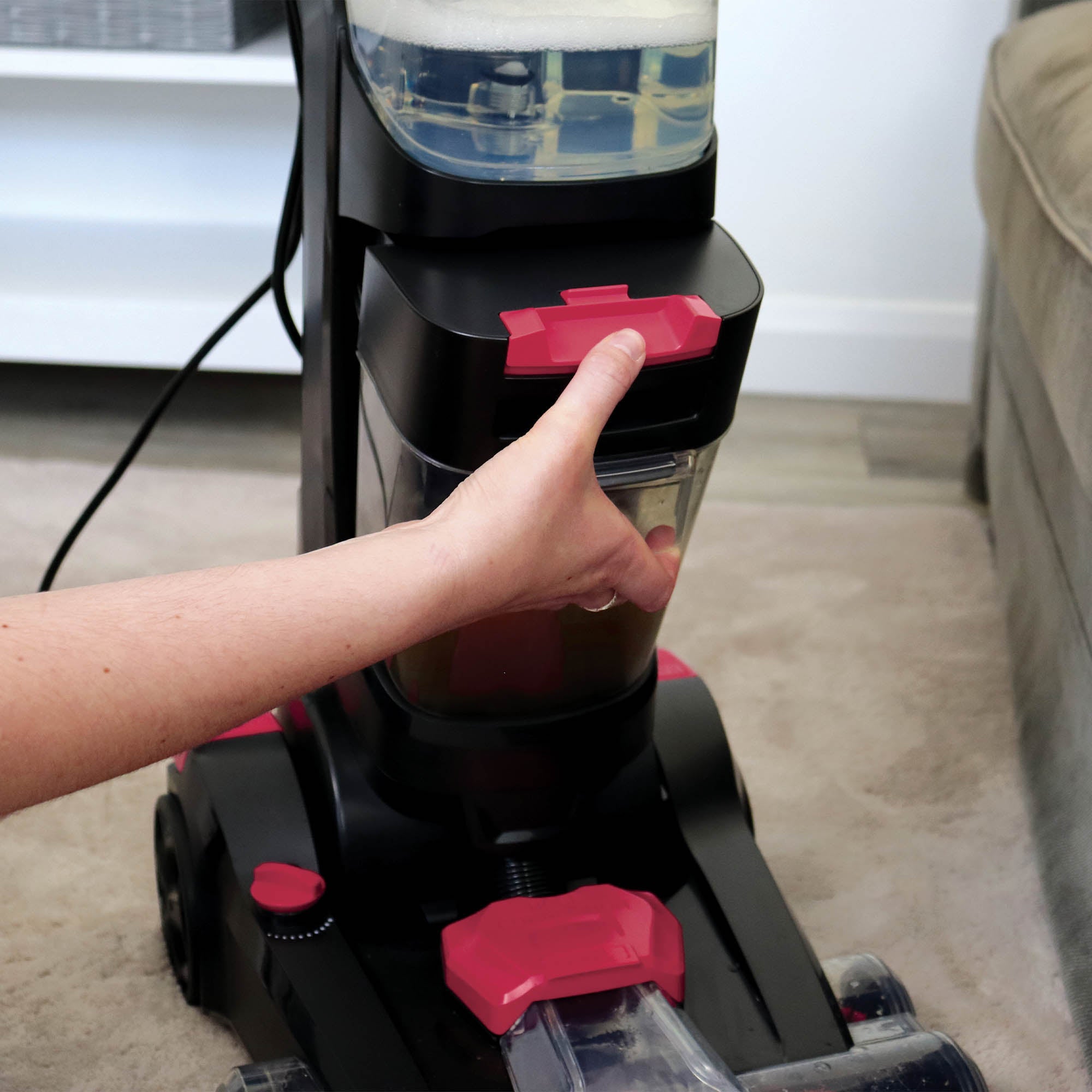 A hand removes the water tank from the HYDROC2 Carpet & Upholstery Cleaner, which has black and pink accents, powerful suction, and visible water in the tank. It sits on a beige carpet near a sofa.